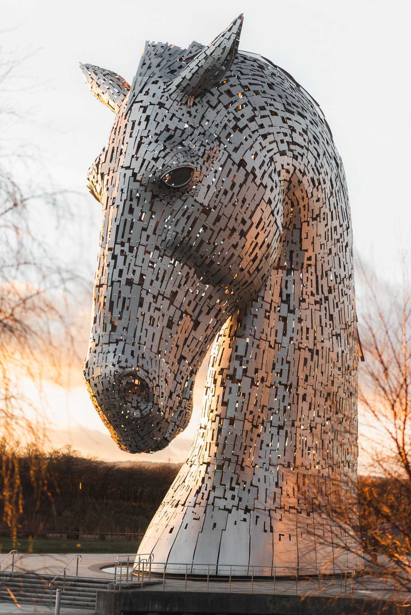 The Kelpies, two large metallic horse-head sculptures, are located outdoors during sunset with trees in the background.