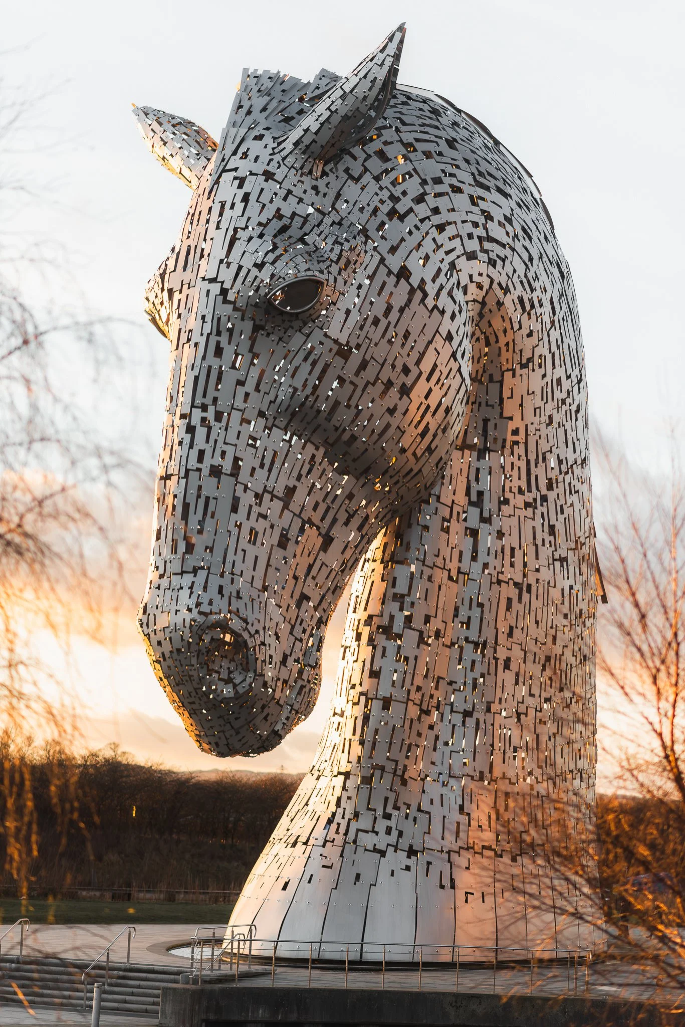 The Kelpies, two large metallic horse-head sculptures, are located outdoors during sunset with trees in the background.