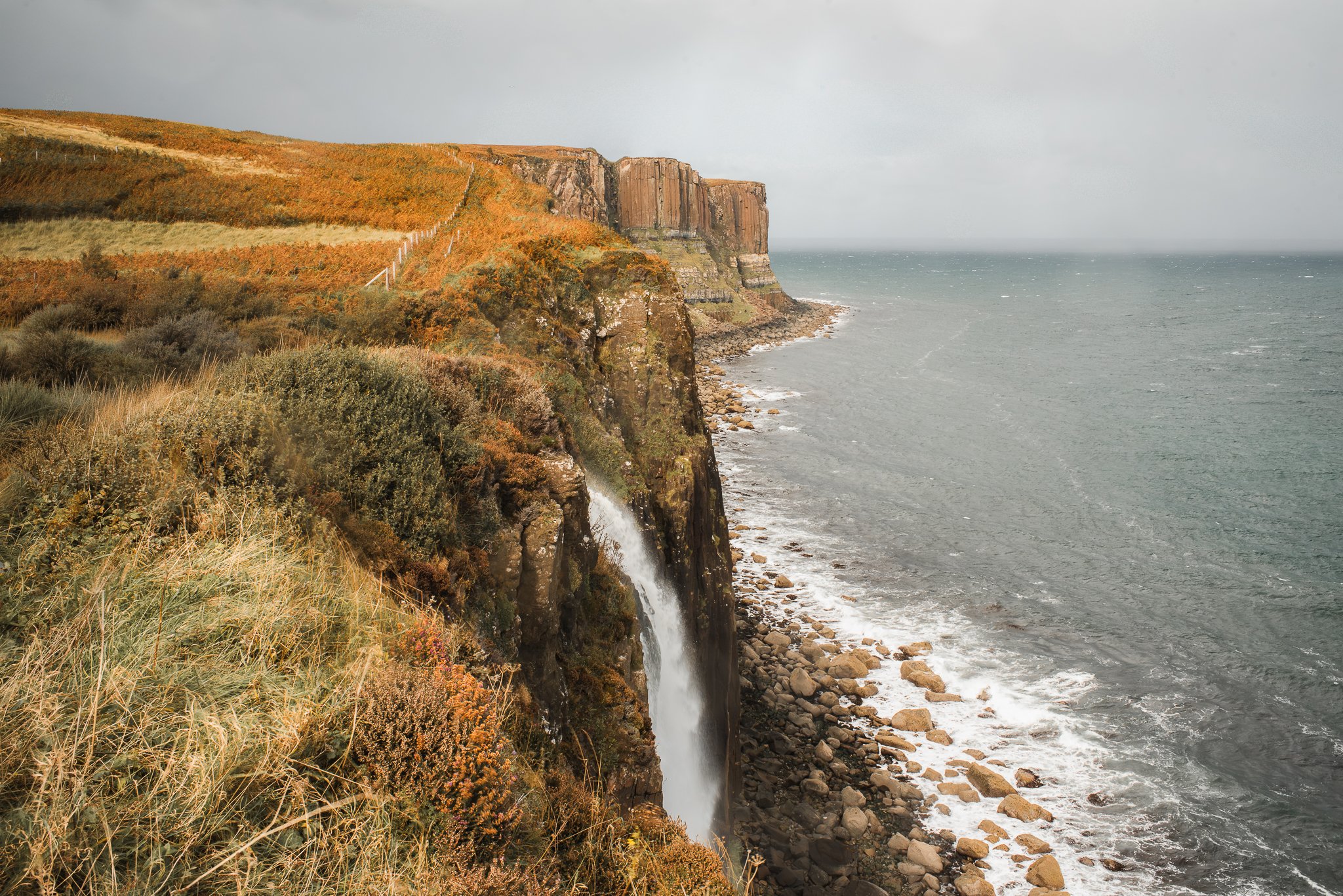 Mealt Falls and Kilt Rock on the edge of the Isle of Skye showing a coastline with a waterfall cascading down, with rocky shore below and overcast sky.