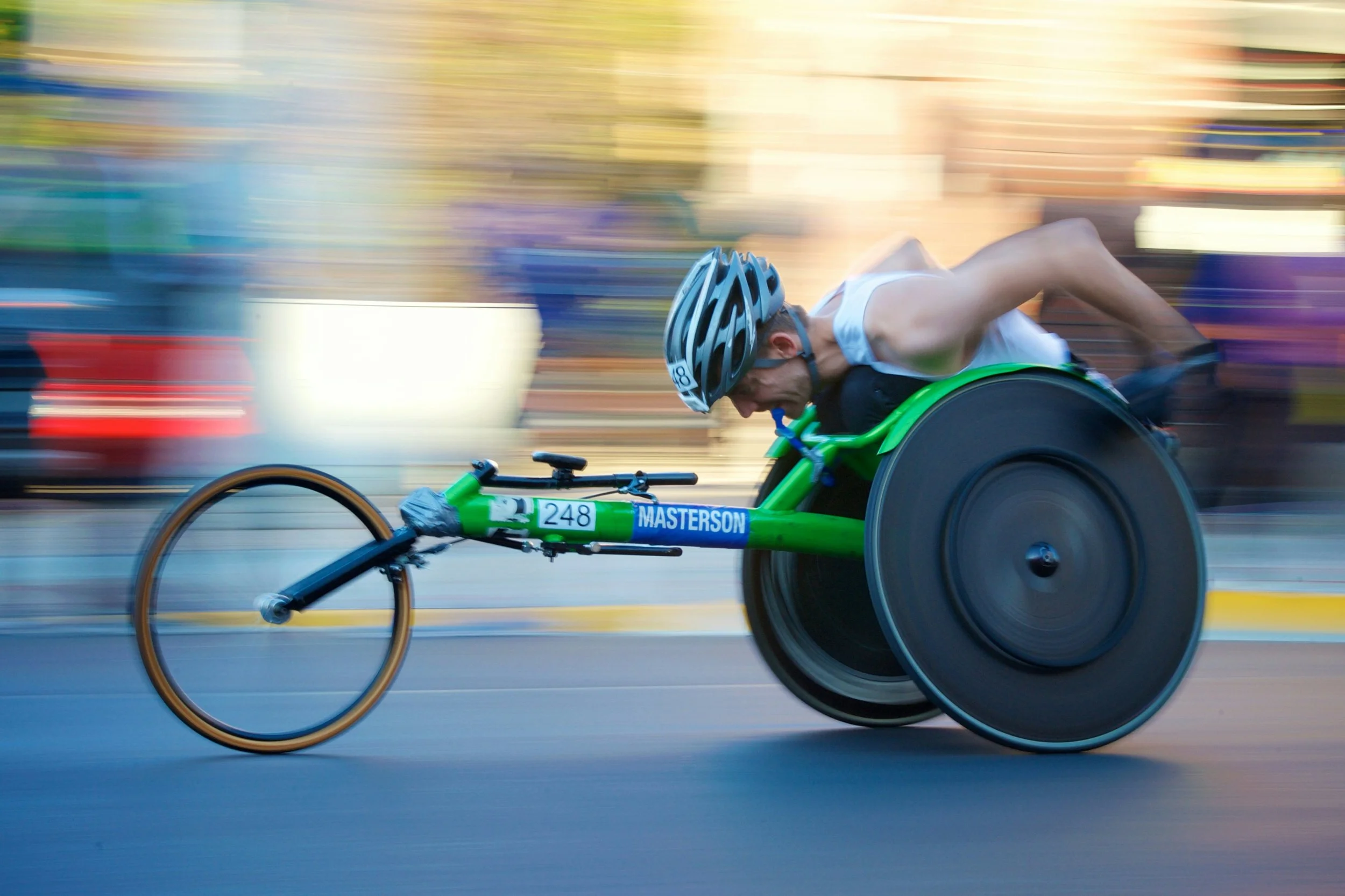A male athlete racing in a wheelchair on a city street, wearing a helmet and white tank top, with blurred background indicating motion.