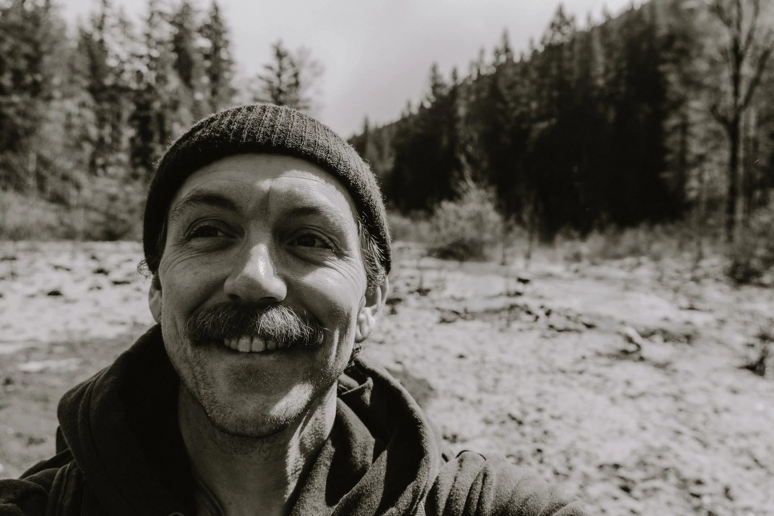 A smiling man with a mustache, wearing a beanie and jacket, taking a selfie outdoors in a forested area.