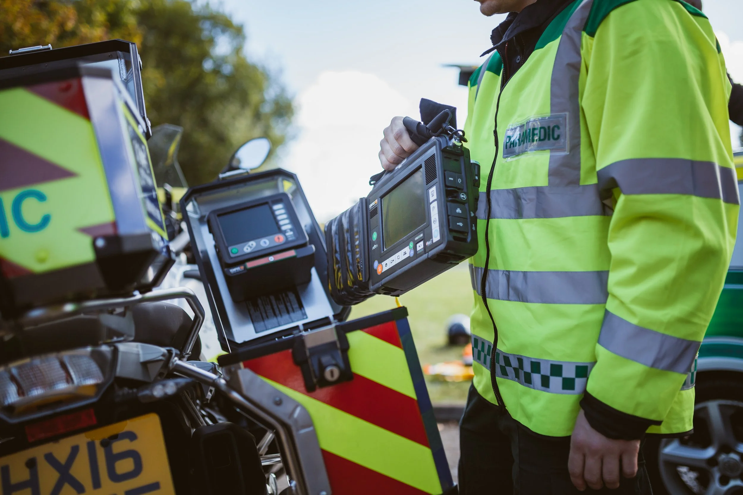 Emergency medical responder in a yellow high-visibility jacket holds a portable monitor on a motorcycle with police markings.