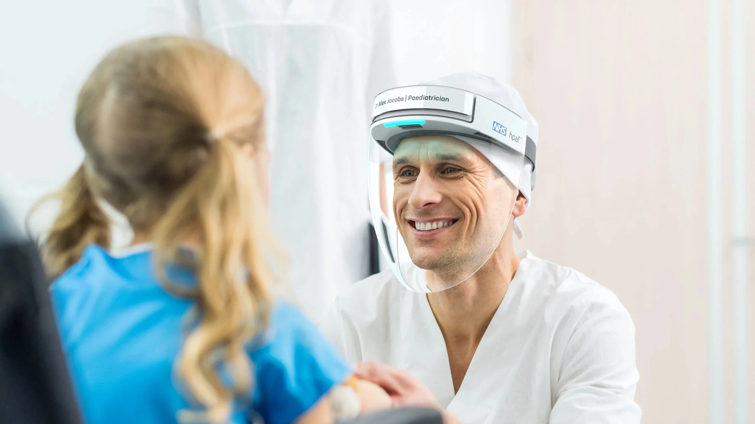 A male healthcare professional wearing a face shield and white medical scrubs smiling at a young girl with long blonde hair, sitting in a hospital or clinic setting.