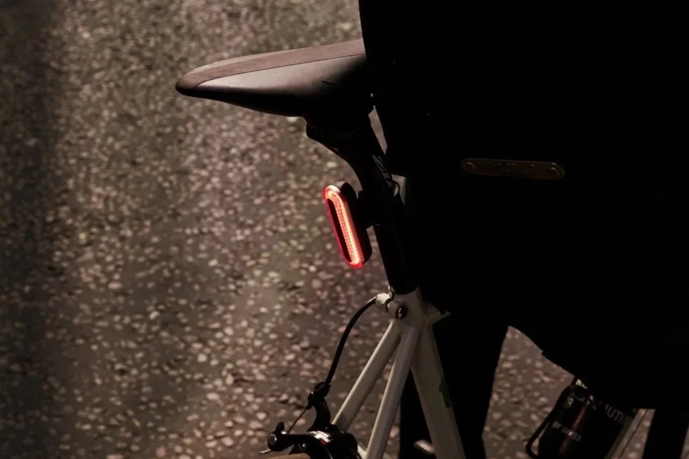 Close-up of the back of a bicycle with an illuminated red tail light, on a dark textured road surface.