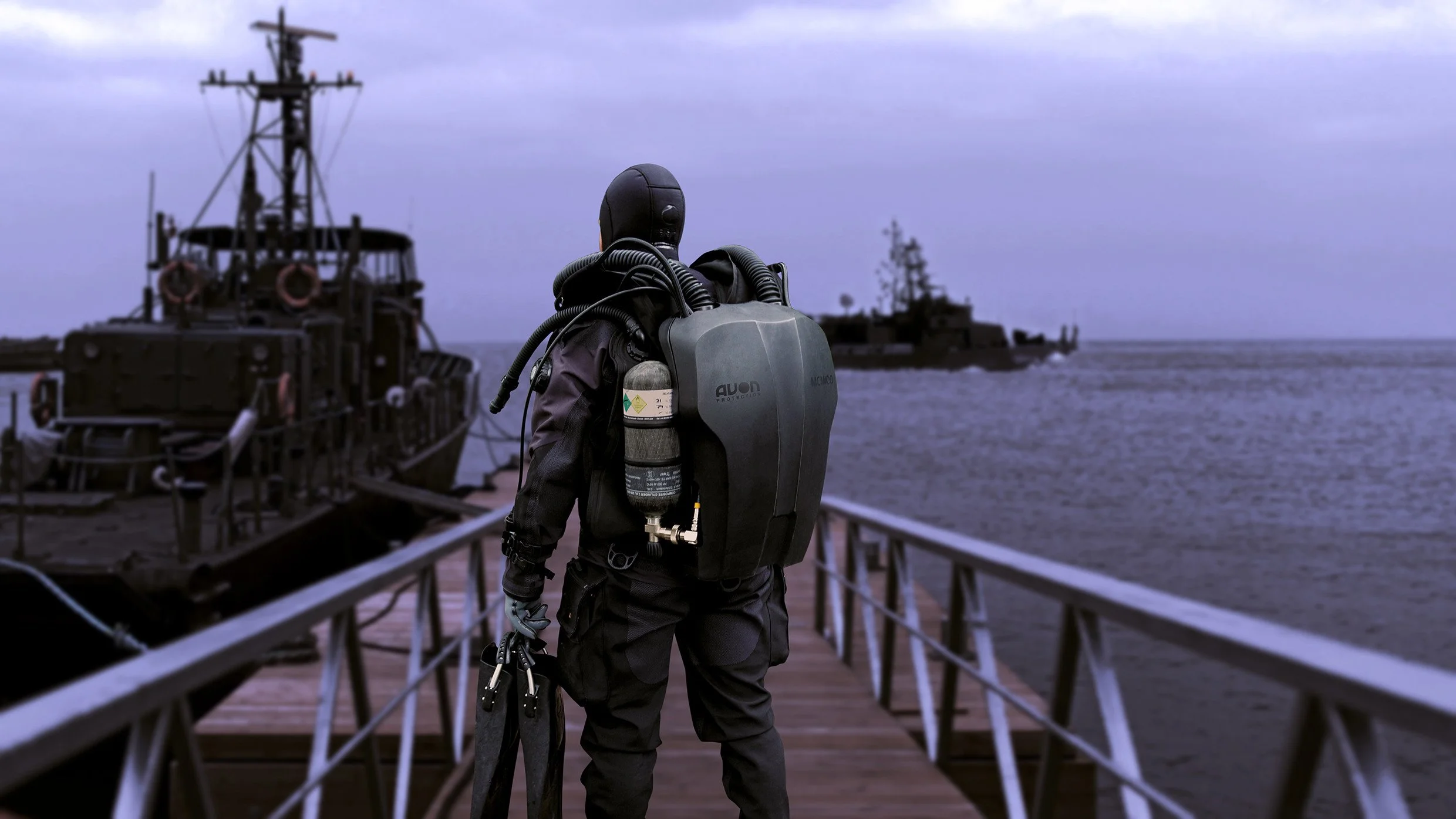 Person in black diving gear with a rebreather tank on their back on a dock, facing towards a naval ship in the water, with another ship in the background, under a cloudy sky.