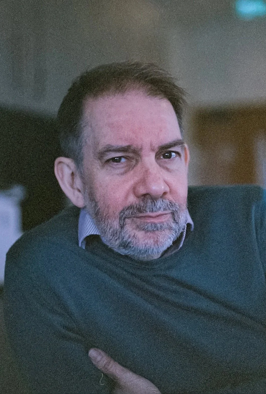 A middle-aged man with short dark hair and a gray beard, wearing a collared shirt and a dark sweater, looking directly at the camera with a calm expression.