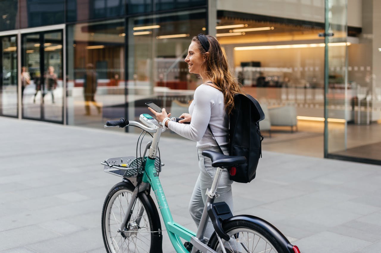 A woman with long brown hair, wearing a white long-sleeve shirt and a black headband, stands next to a teal bicycle outside a modern glass building. She is smiling and looking up from her smartphone, with a black backpack on her back.