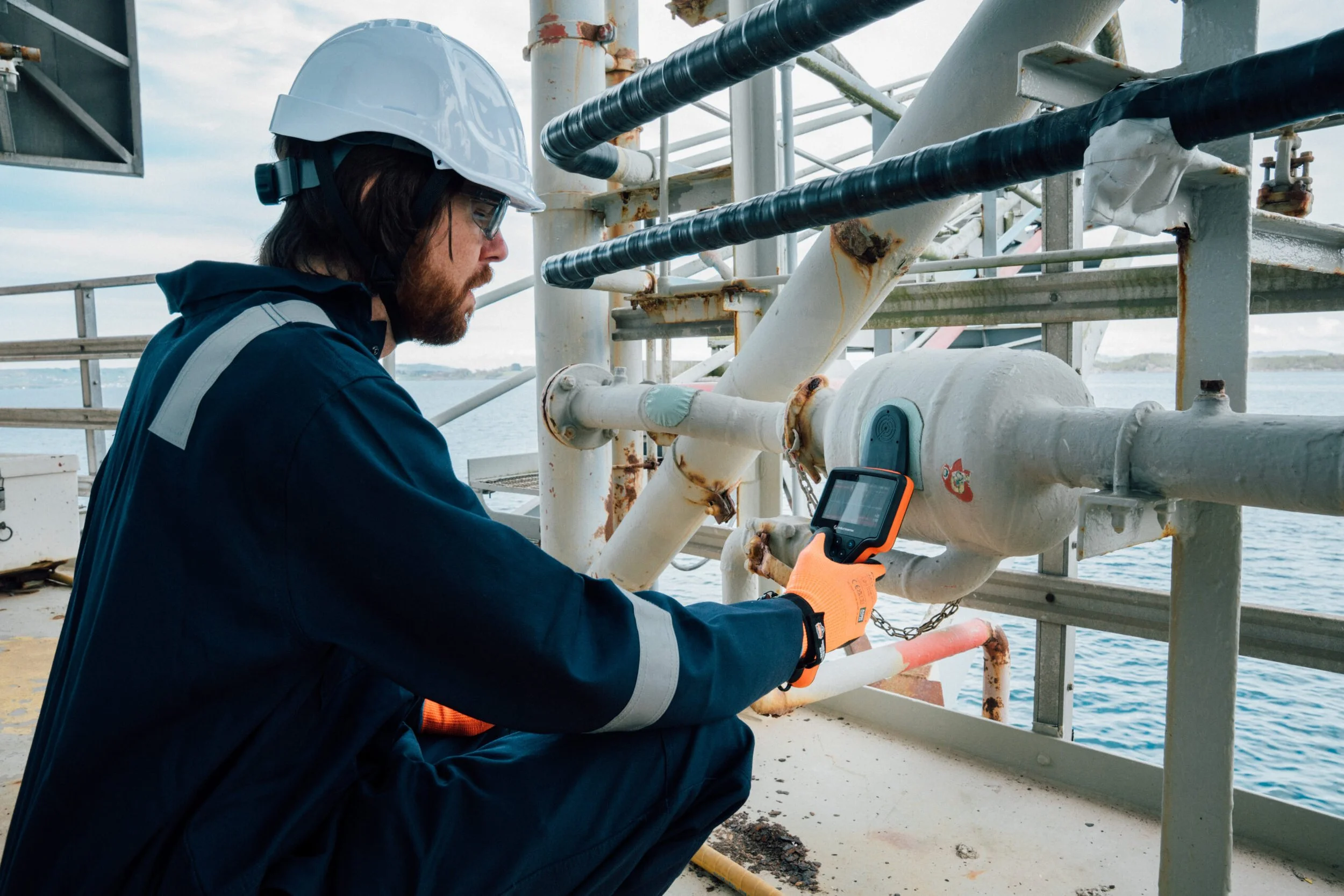 A man wearing a hard hat, safety glasses, an orange glove, and a dark blue coveralls kneels on a ship's deck, inspecting a pipe system with a handheld device, overlooking the ocean.