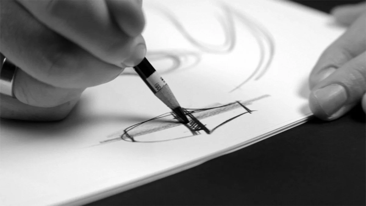 A person drawing a sketch of a revolved form on white paper using a black pencil.