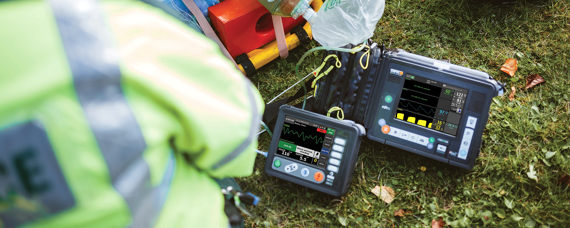 Medical personnel wearing neon yellow safety vests using portable heart monitor devices outdoors on grass, connected to medical supplies and equipment for emergency response.