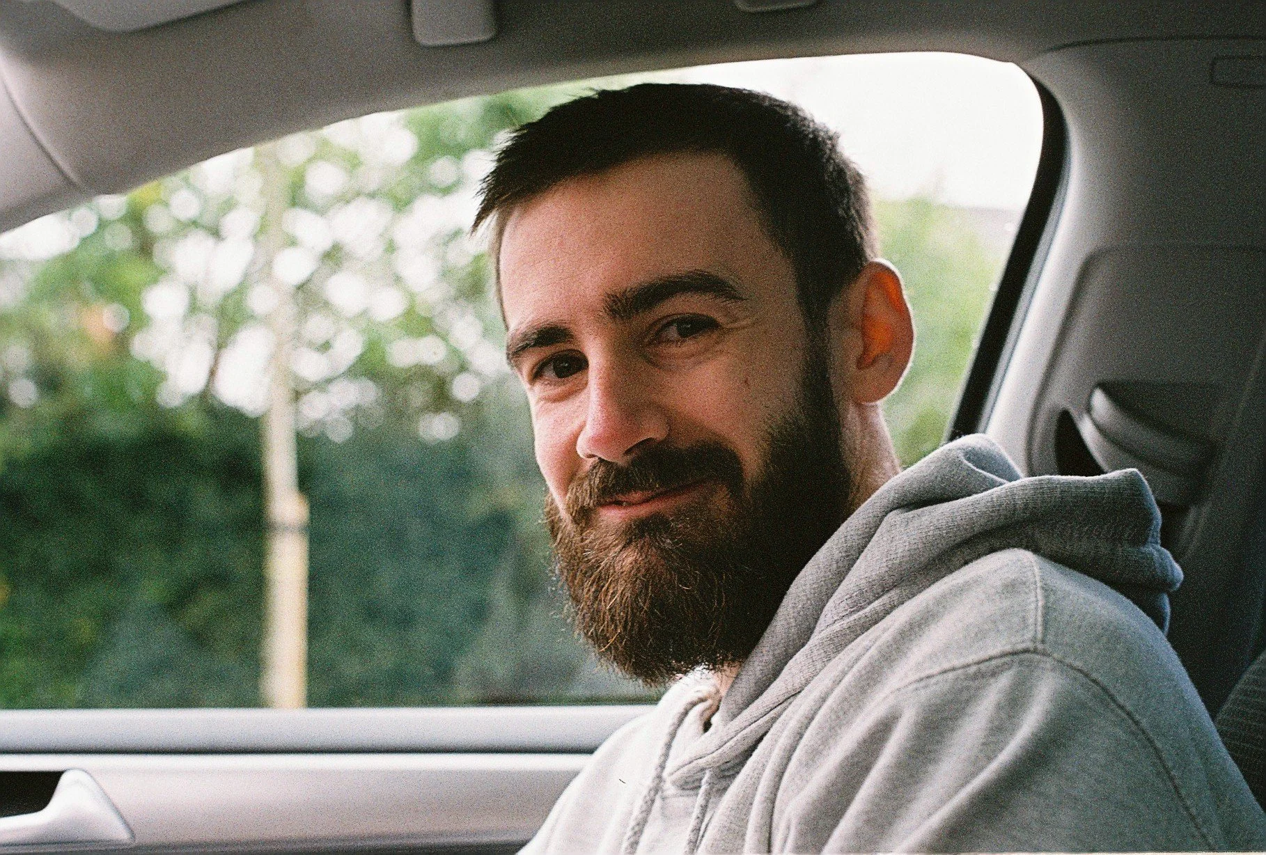 A man with a beard and short dark hair sitting inside a vehicle, looking at the camera with a slight smile, with blurred greenery visible outside the window.