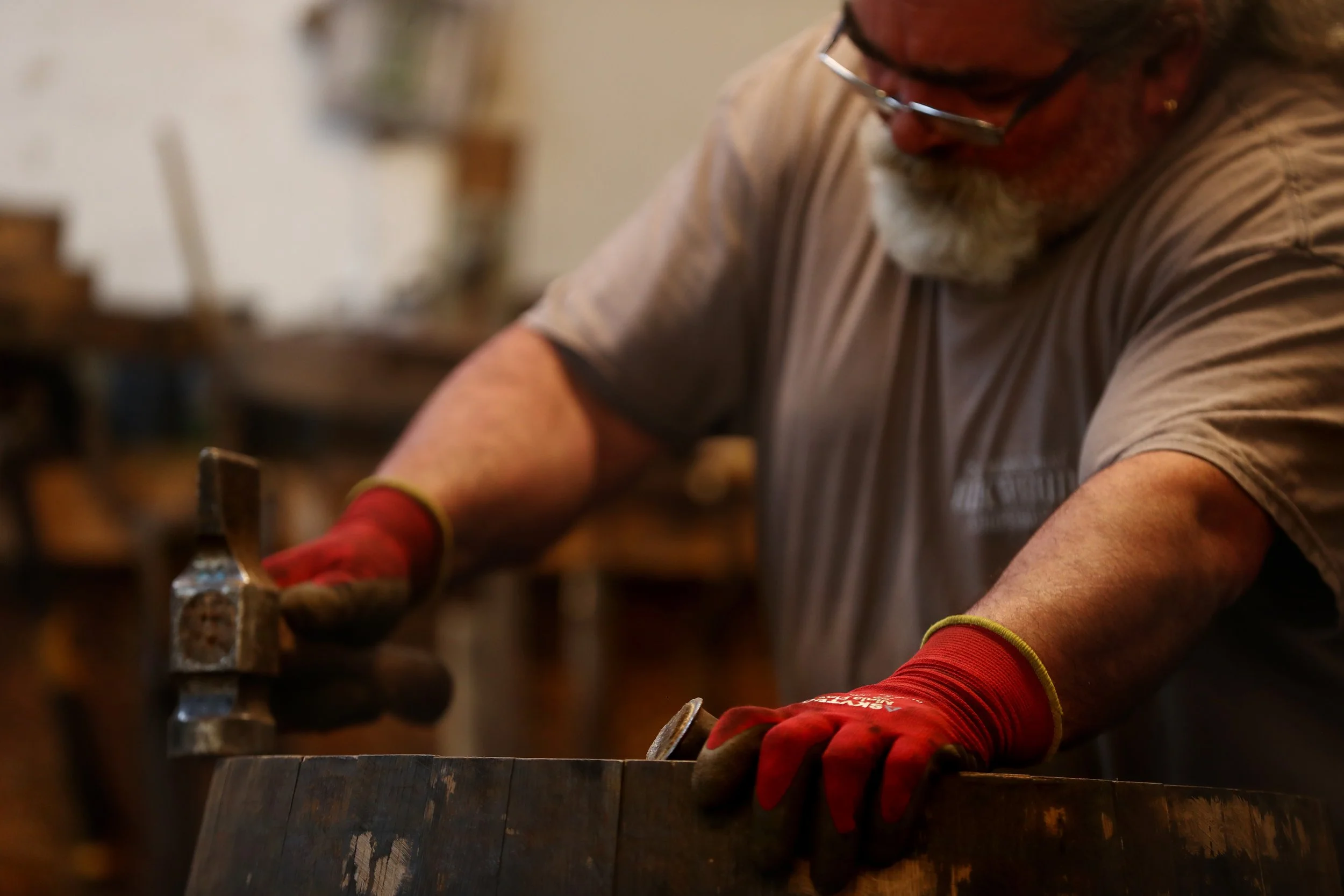 A cooper wearing glasses, a beige t-shirt, red gloves, and a beard is working on a wooden barrel in a workshop.