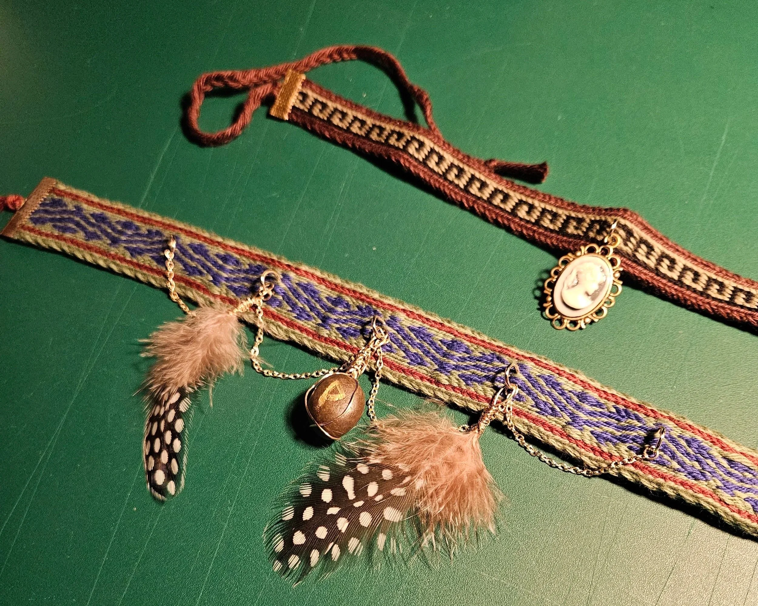 Deux colliers en tissage aux cartes, un avec un médaillon camé de style ancien et un autre avec des plumes de faisan, des perles, et une pierre brute gravée d'une rune.