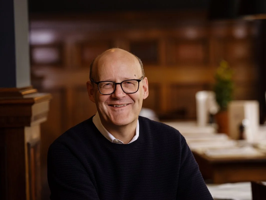 Ein lächelnder Mann mit Brille in einem dunklen Pullover in einem gemütlichen Raum mit Holzvertäfelung im Hintergrund.