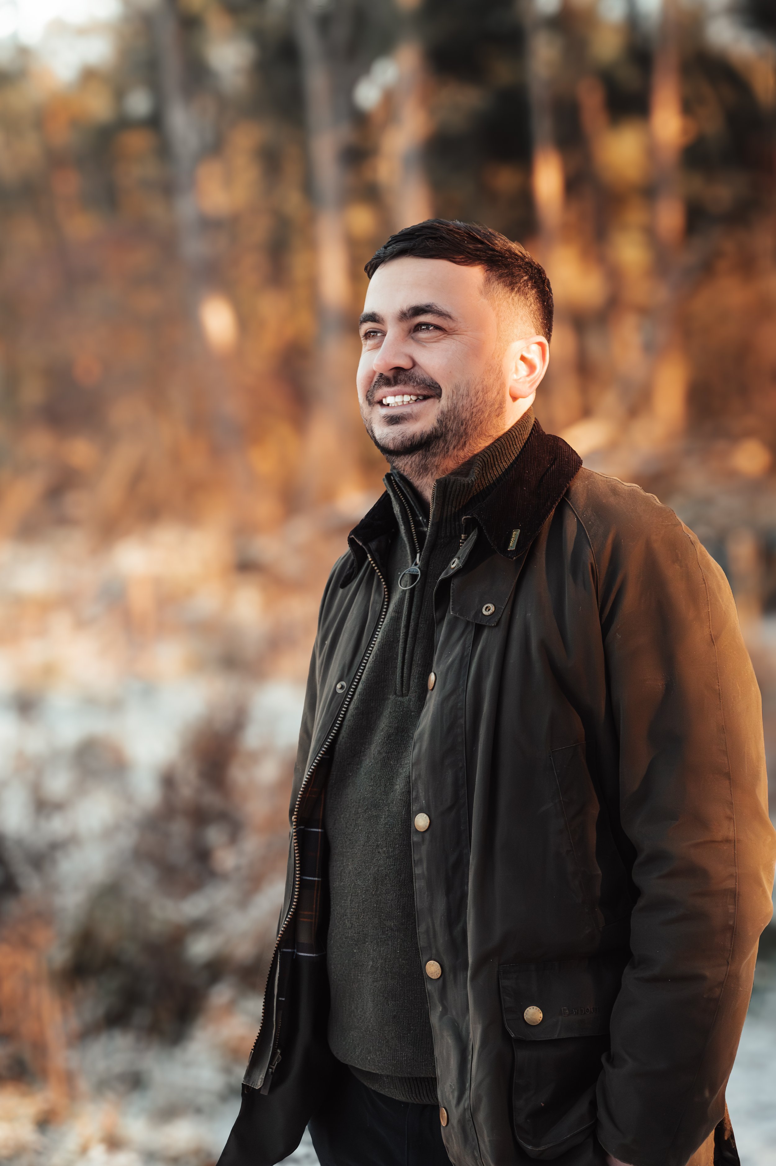 Man standing outdoors during sunset with a smile, dressed in black jacket and sweater, background featuring autumn trees and rocky terrain.