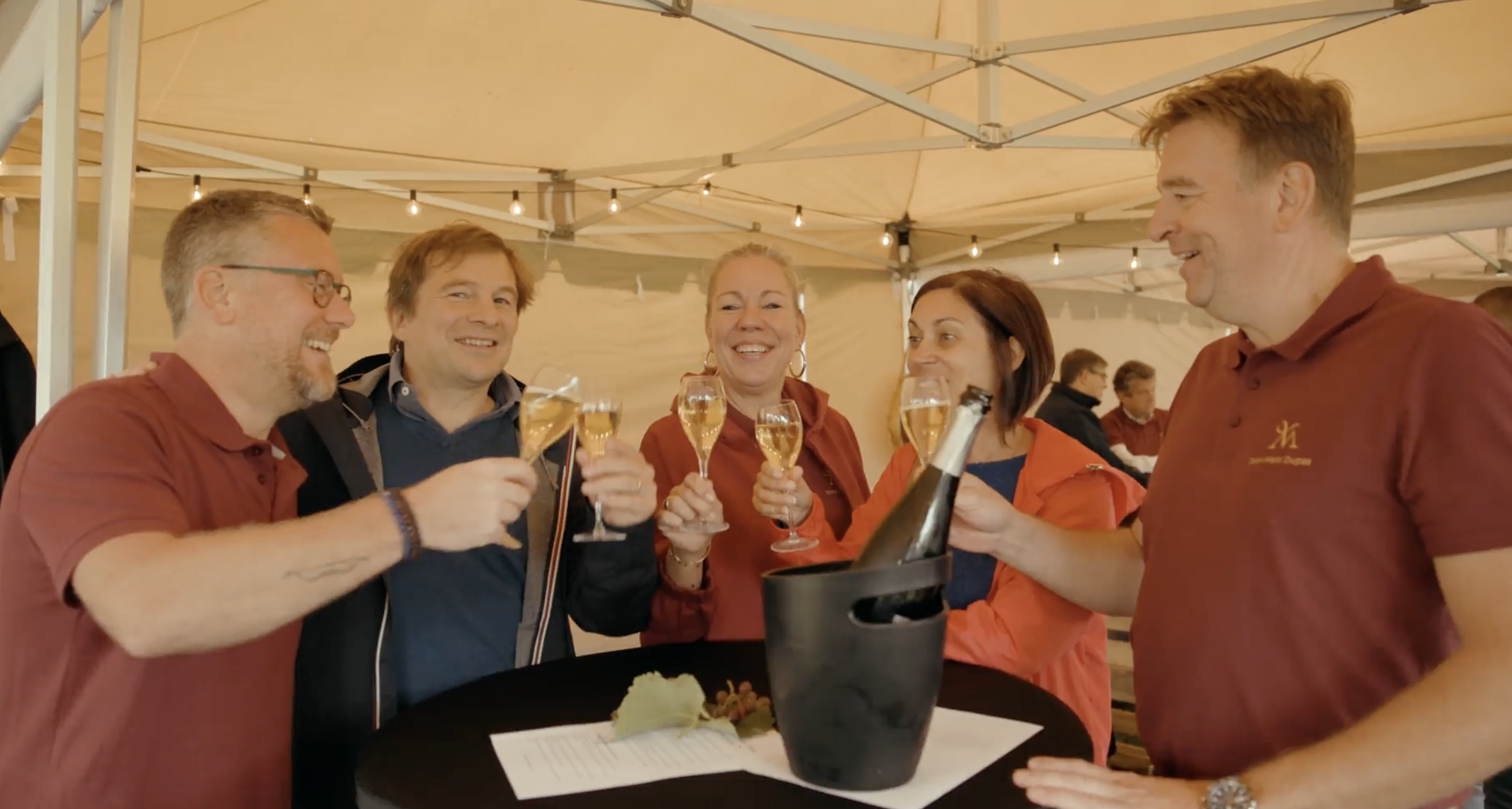 Groupe de cinq personnes souriantes lors d'une célébration avec des verres de champagne, sous une tente avec des lumières suspendues.