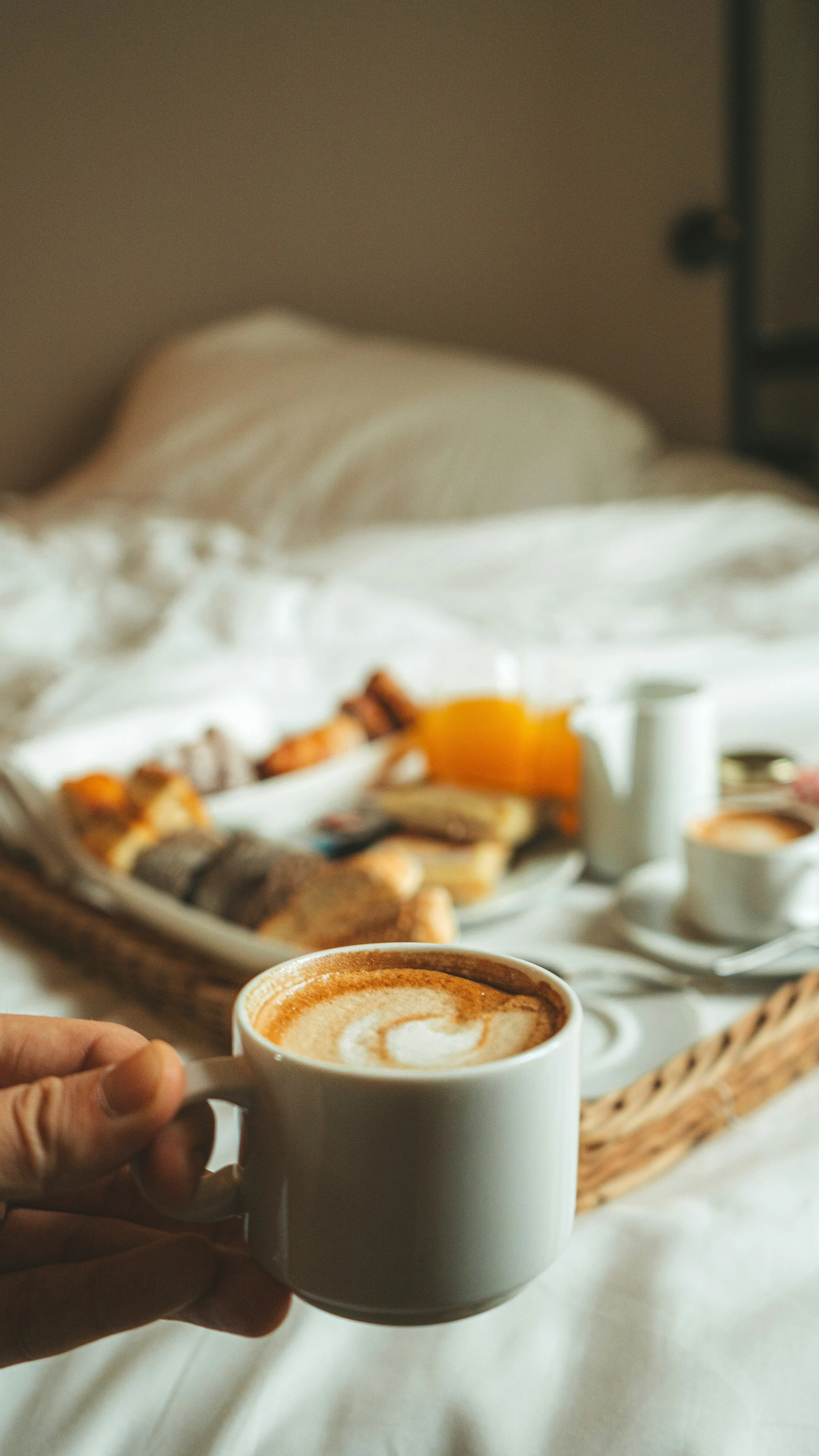 A person holding a white mug of coffee with foam art, in a cozy setting with breakfast items like croissants, juice, and a cup of tea on a tray in the background.