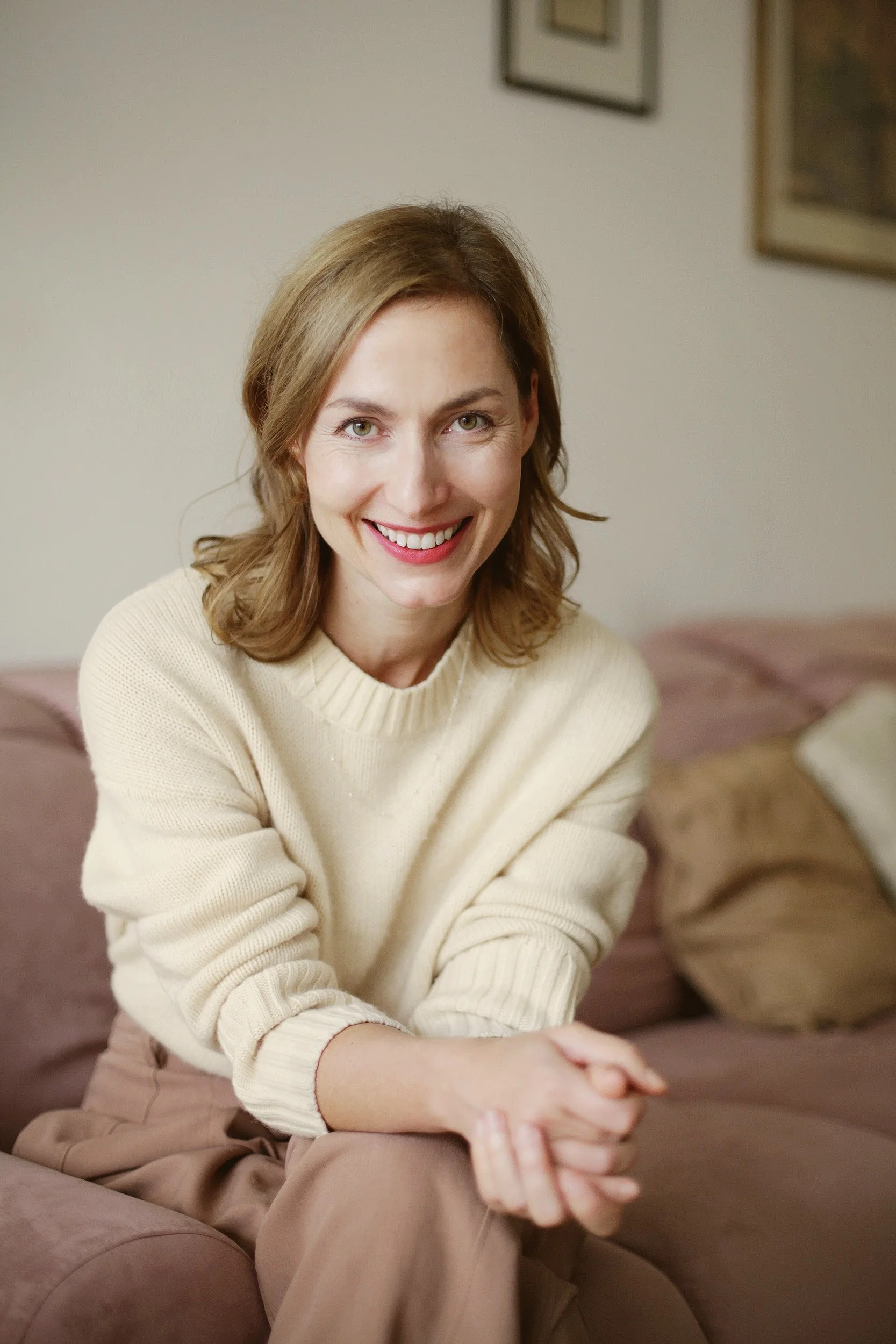 A woman with shoulder-length light brown hair, wearing a cream-colored sweater, sitting on a pink sofa with beige and white pillows, smiling at the camera.