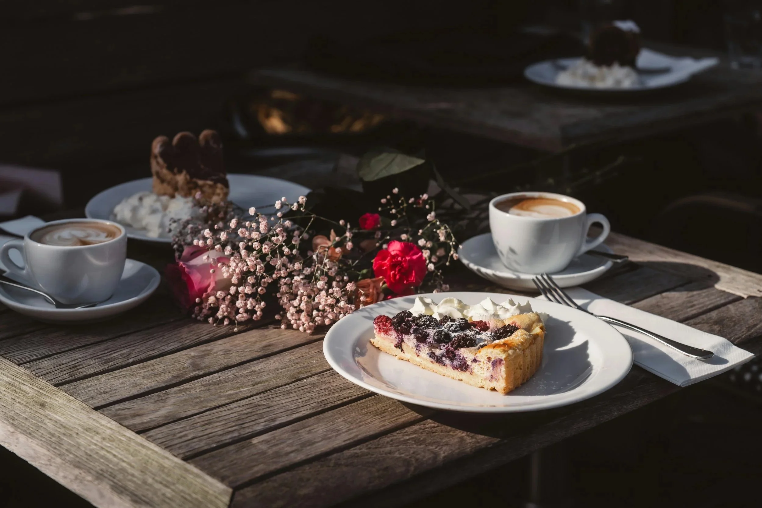 A wooden outdoor table set with a slice of mixed berry tart with whipped cream, two cups of coffee, a plate with ice cream and a cookie, a bouquet of pink and red flowers, and sunlight casting shadows.