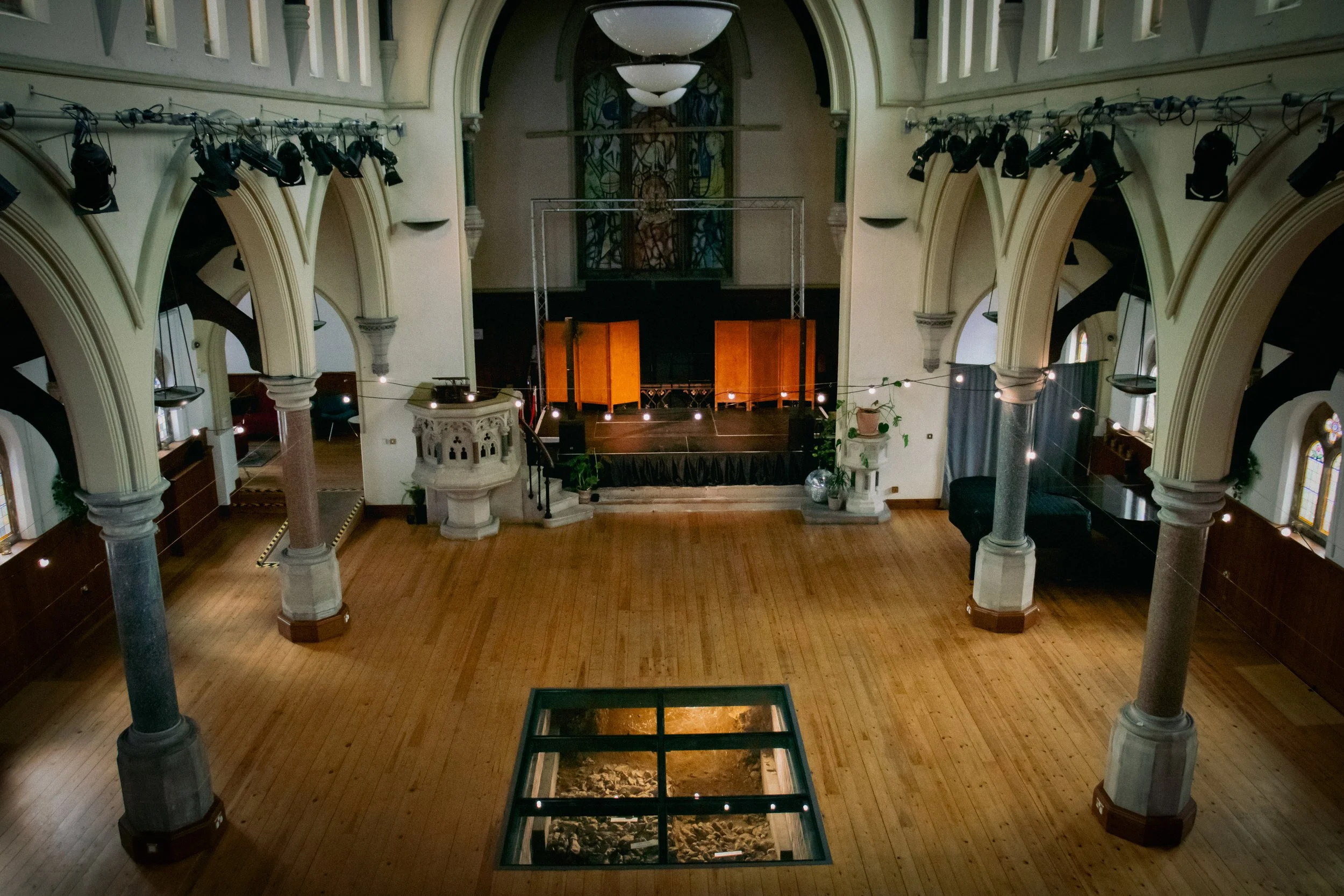 Empty historic church or hall with wooden floors, arched windows, columns, and a stage at the front, decorated with string lights and plants.