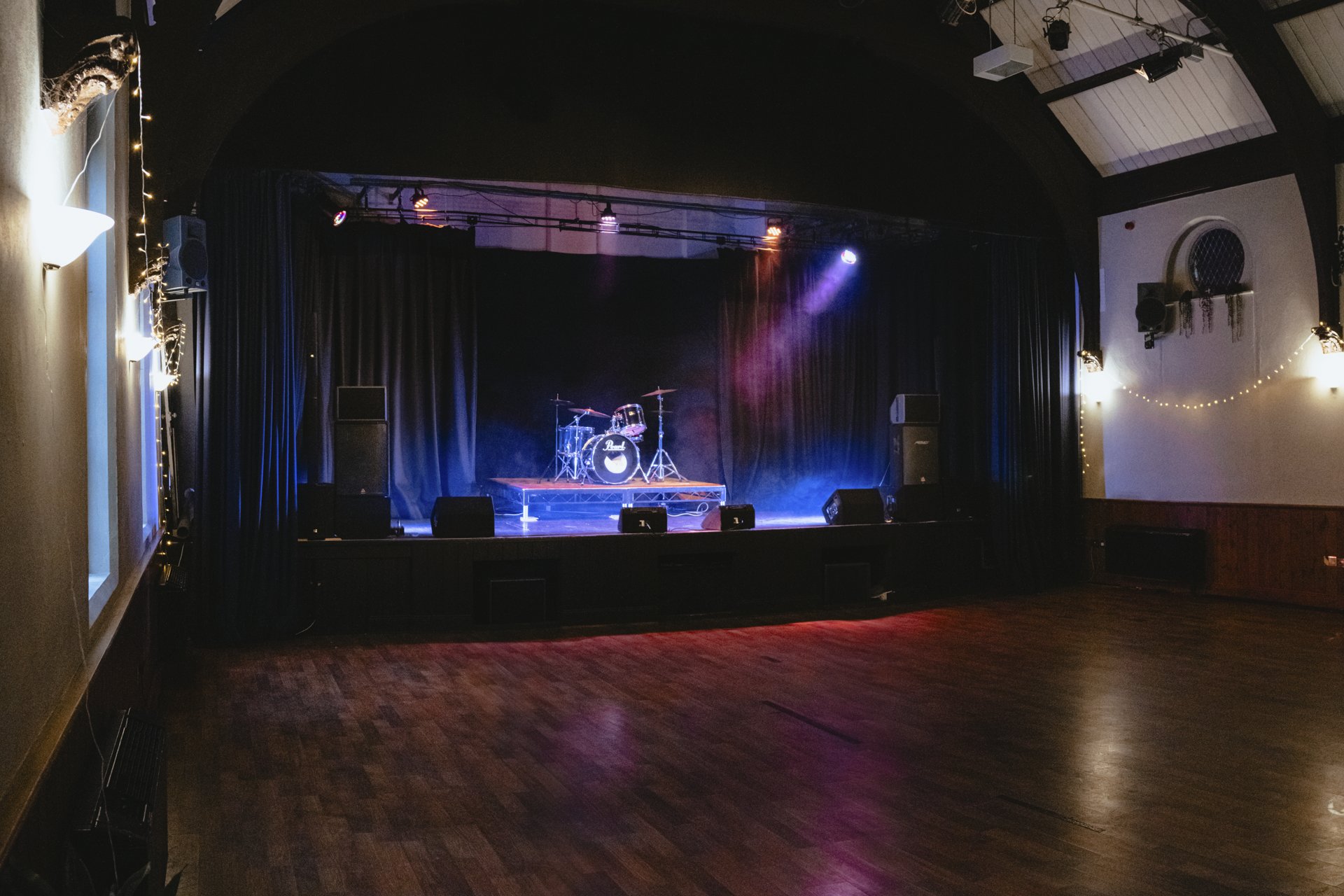 Empty stage with a drum set, surrounded by black curtains, with colorful stage lights overhead, in a music or performance venue.