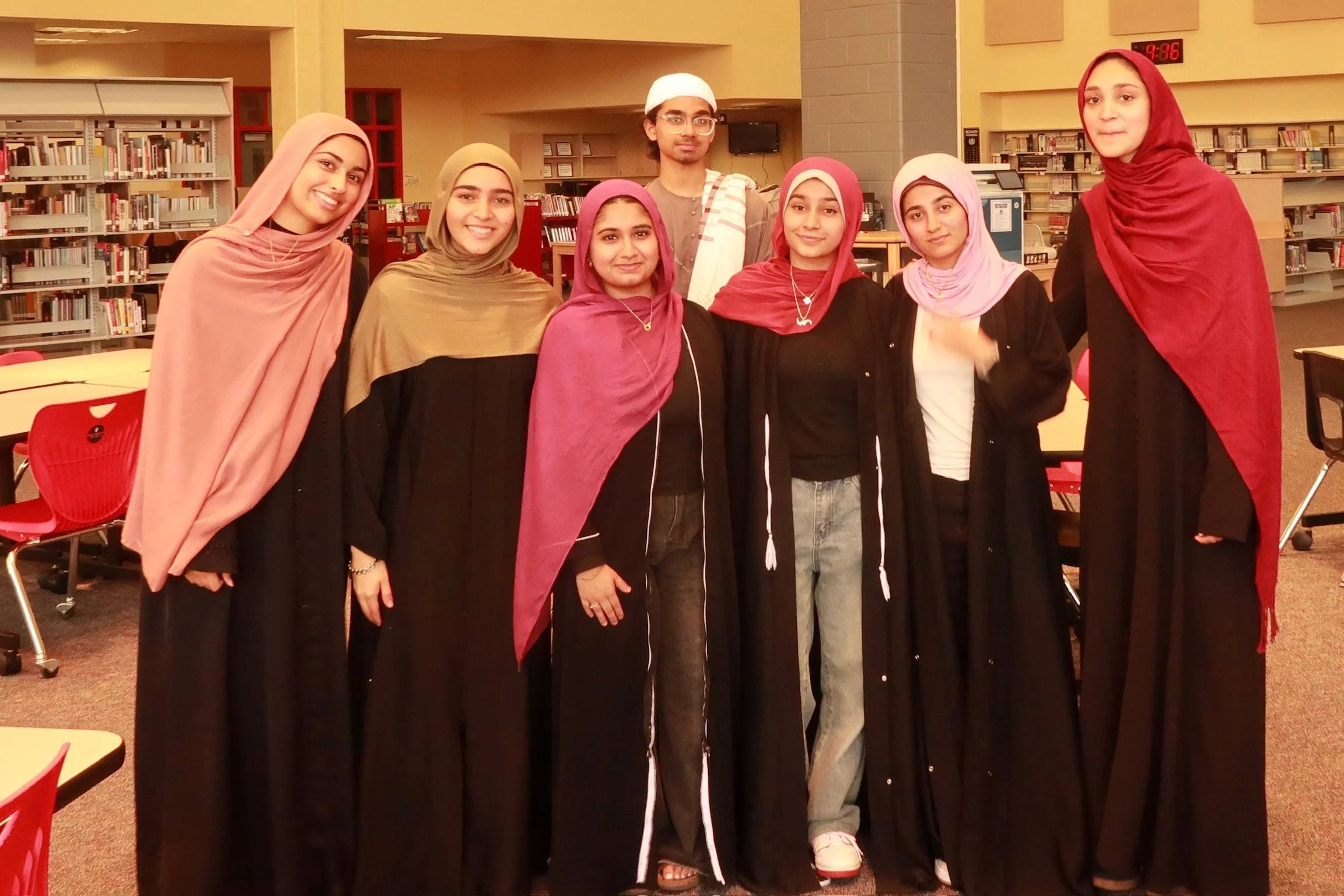Group of young Muslim women and one young man standing together in a library, smiling at the camera.
