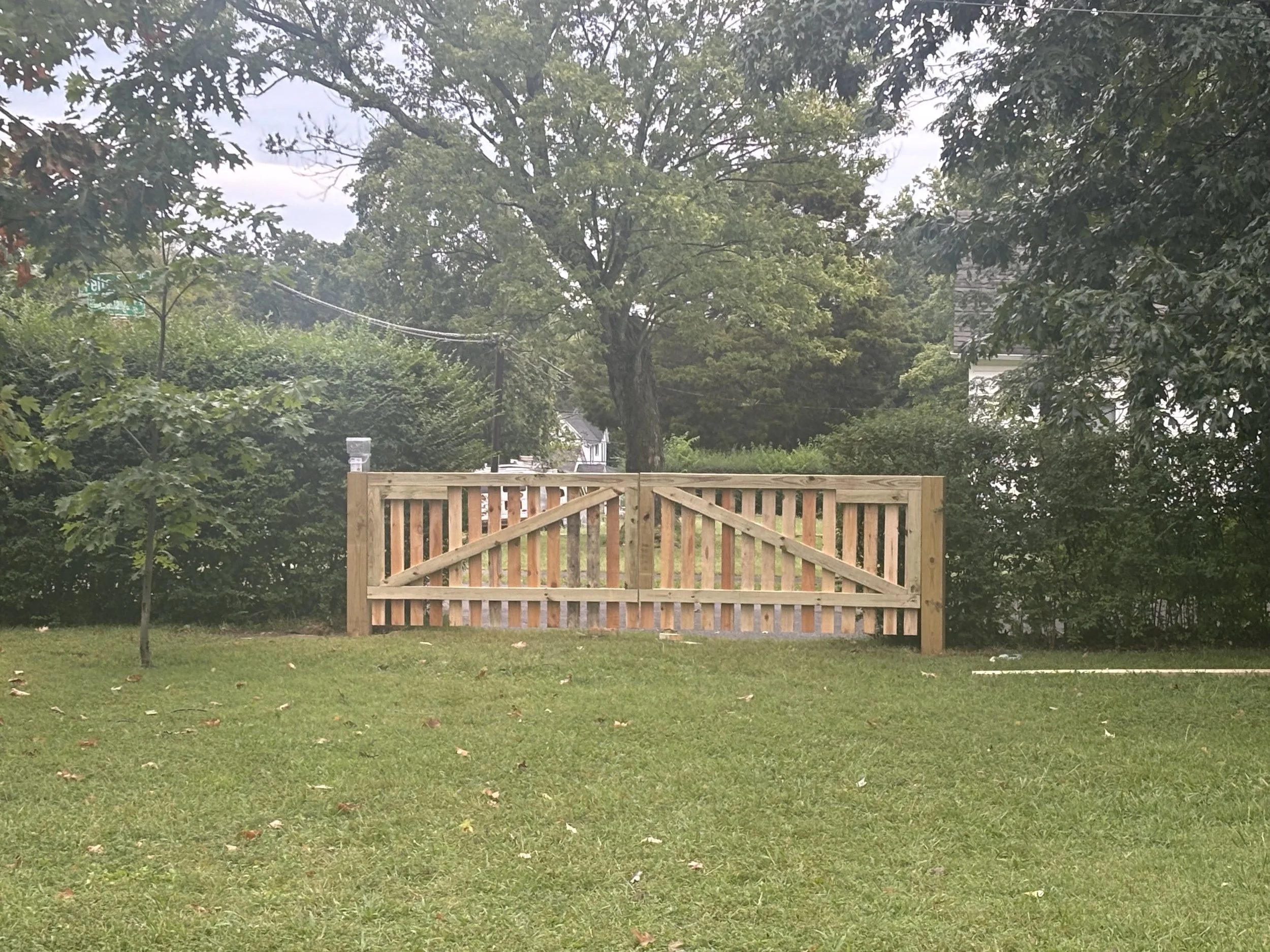 A wooden gate with a diagonal bracing is closed across a grassy yard, with trees and bushes in the background.