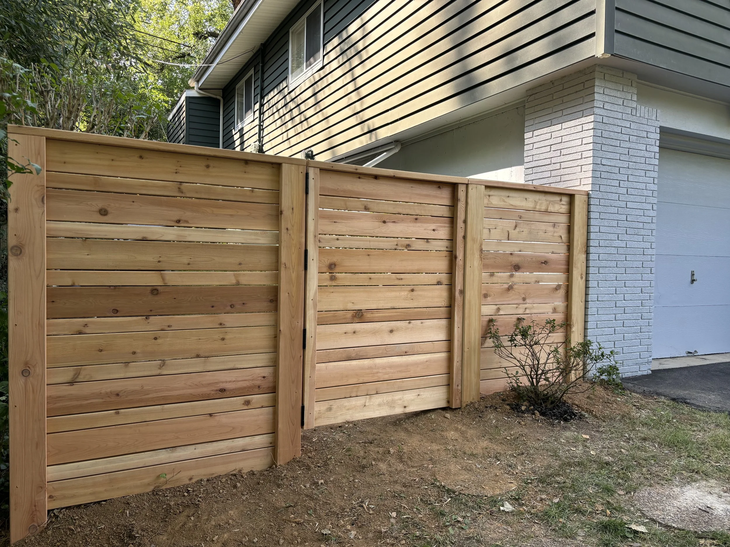 New wooden fence with horizontal slats in front of a house with a white brick wall and a gray garage door.