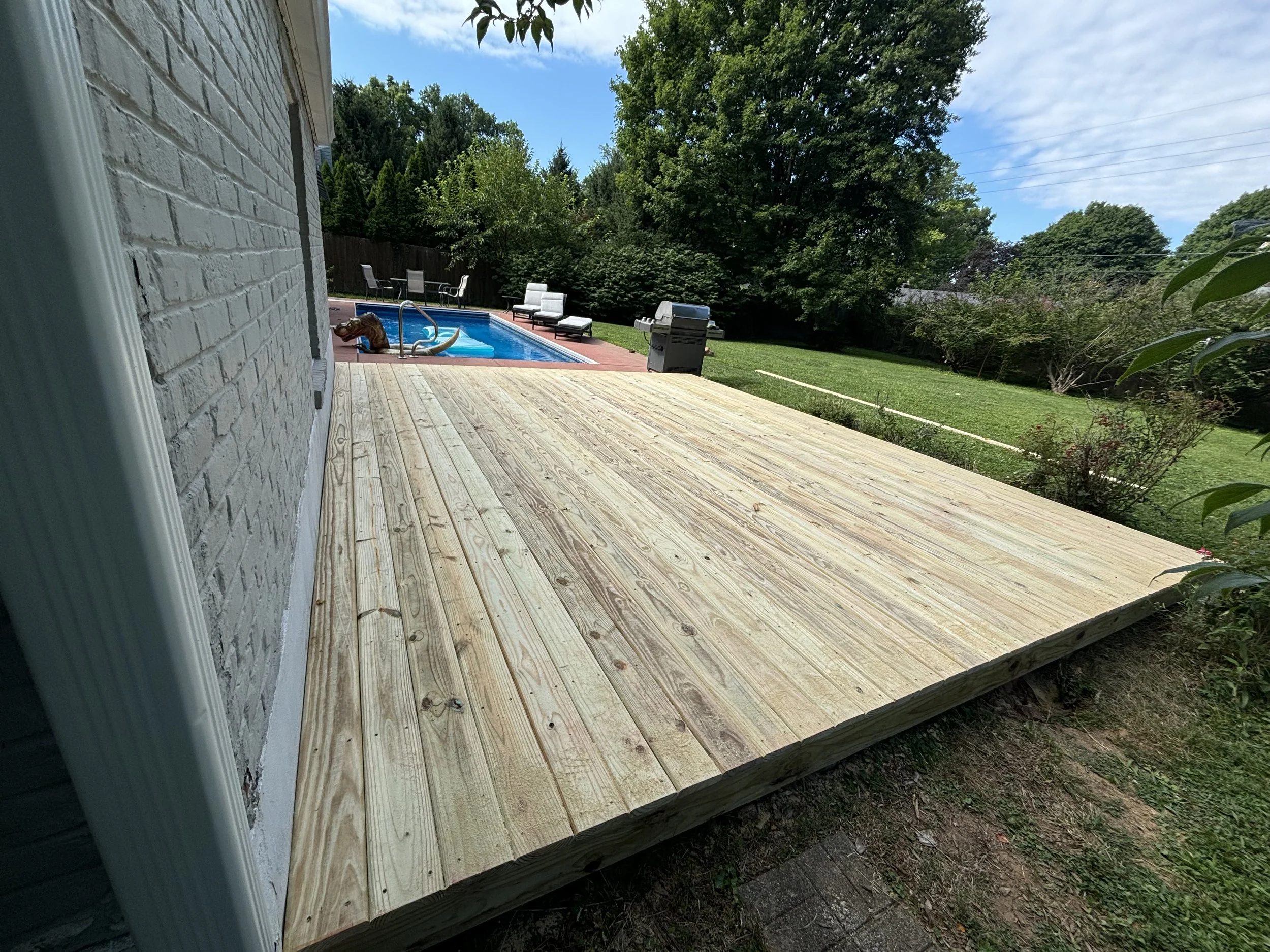 View of a backyard with a newly built wooden deck, pool with a slide, lounge chairs, a barbecue grill, and lush green trees under a blue sky.