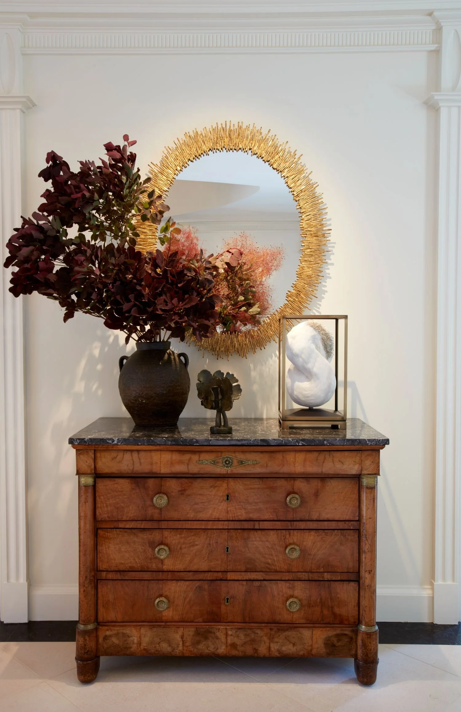 A vintage wooden chest with a dark marble top, decorated with a large dark vase holding purple and pink foliage, a small sculpture, and a modern white sculpture encased in a glass box. Behind it is a round, gold suncatcher mirror on a white wall.