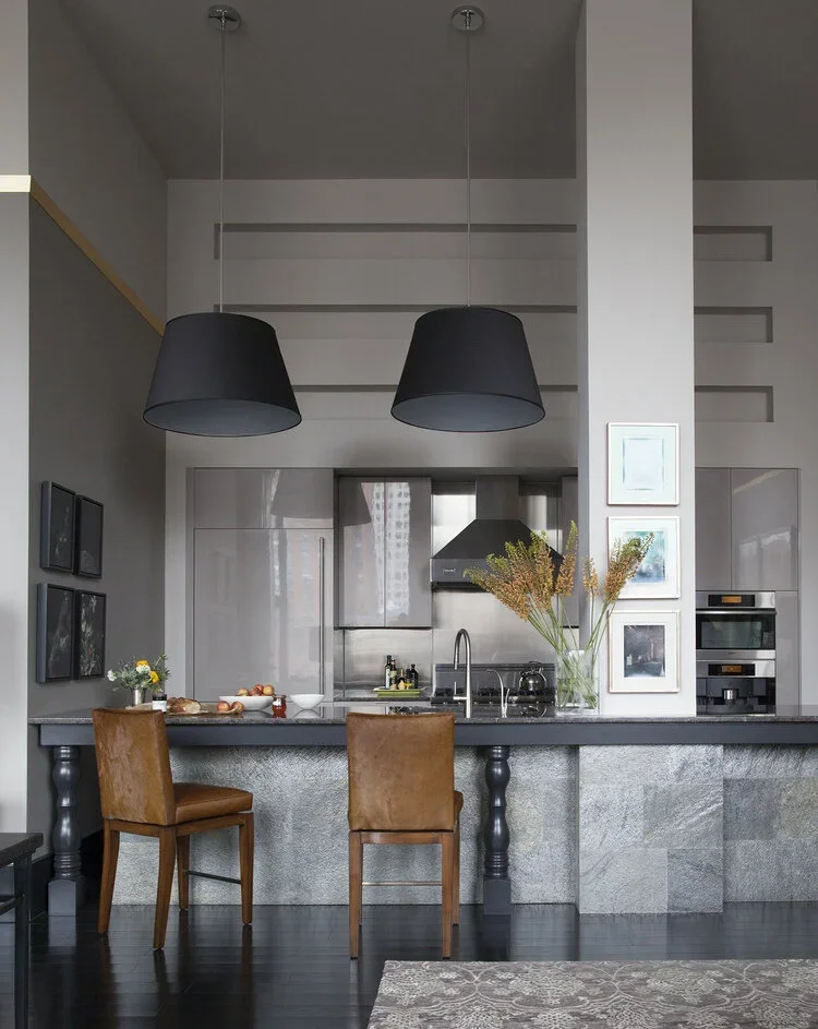 Modern kitchen with gray countertops, two brown chairs at the counter, a vase with flowers, and black pendant lights.