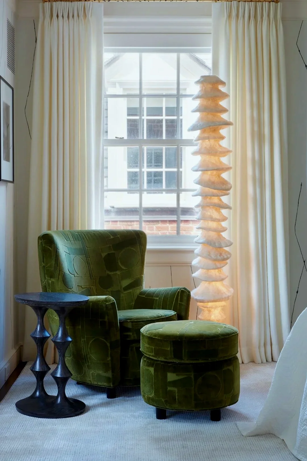 A cozy interior with a green patterned armchair and matching ottoman beside a black side table, positioned in front of a window with cream-colored curtains. A tall, decorative white lamp with a layered, textured design stands next to the window, which shows a glimpse of a neighboring house and brick wall outside.