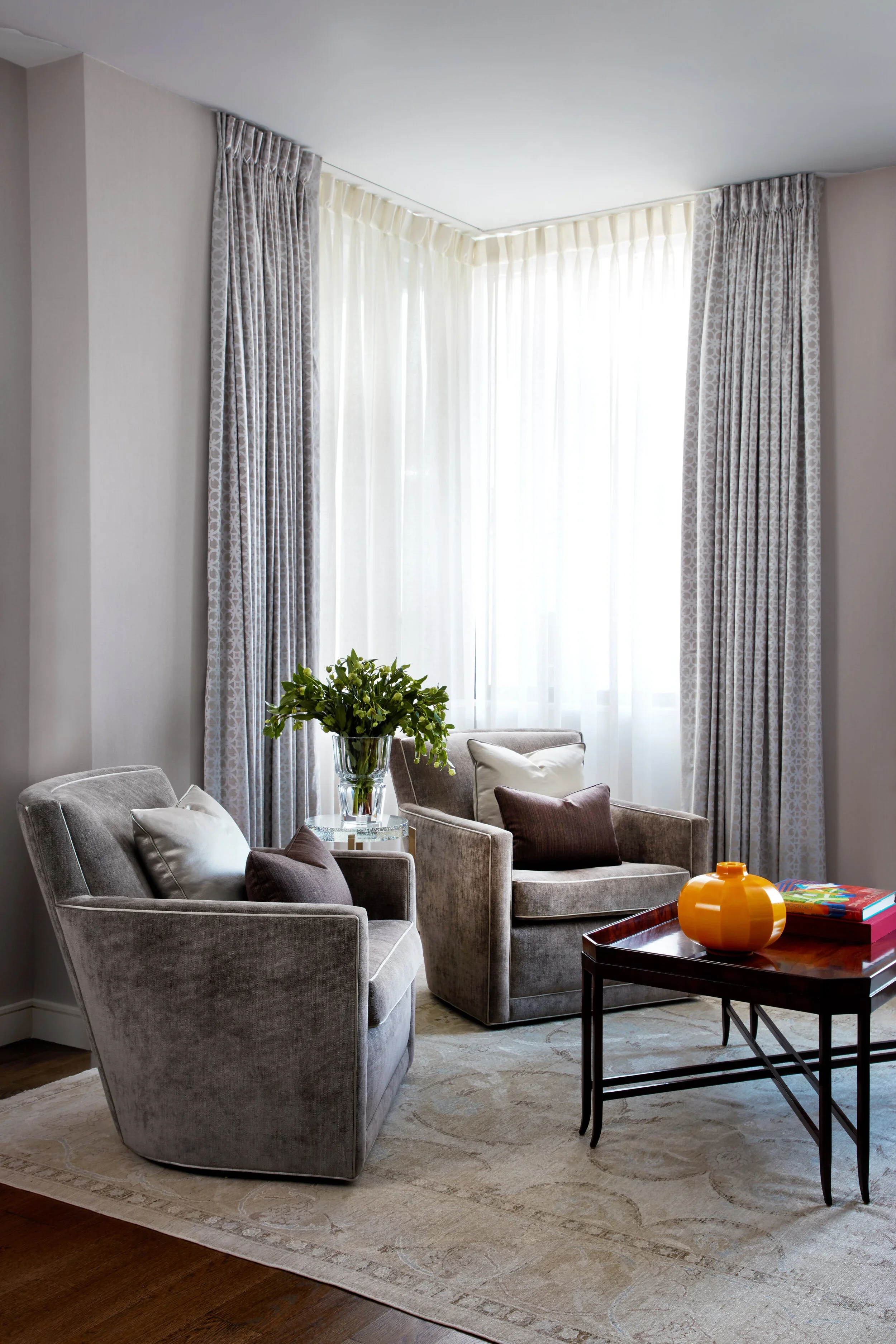 A cozy living room corner with two plush gray armchairs, patterned curtains, a large window with white curtains, a side table with a green plant, and a wooden coffee table with a yellow pumpkin-shaped vase and colorful books.