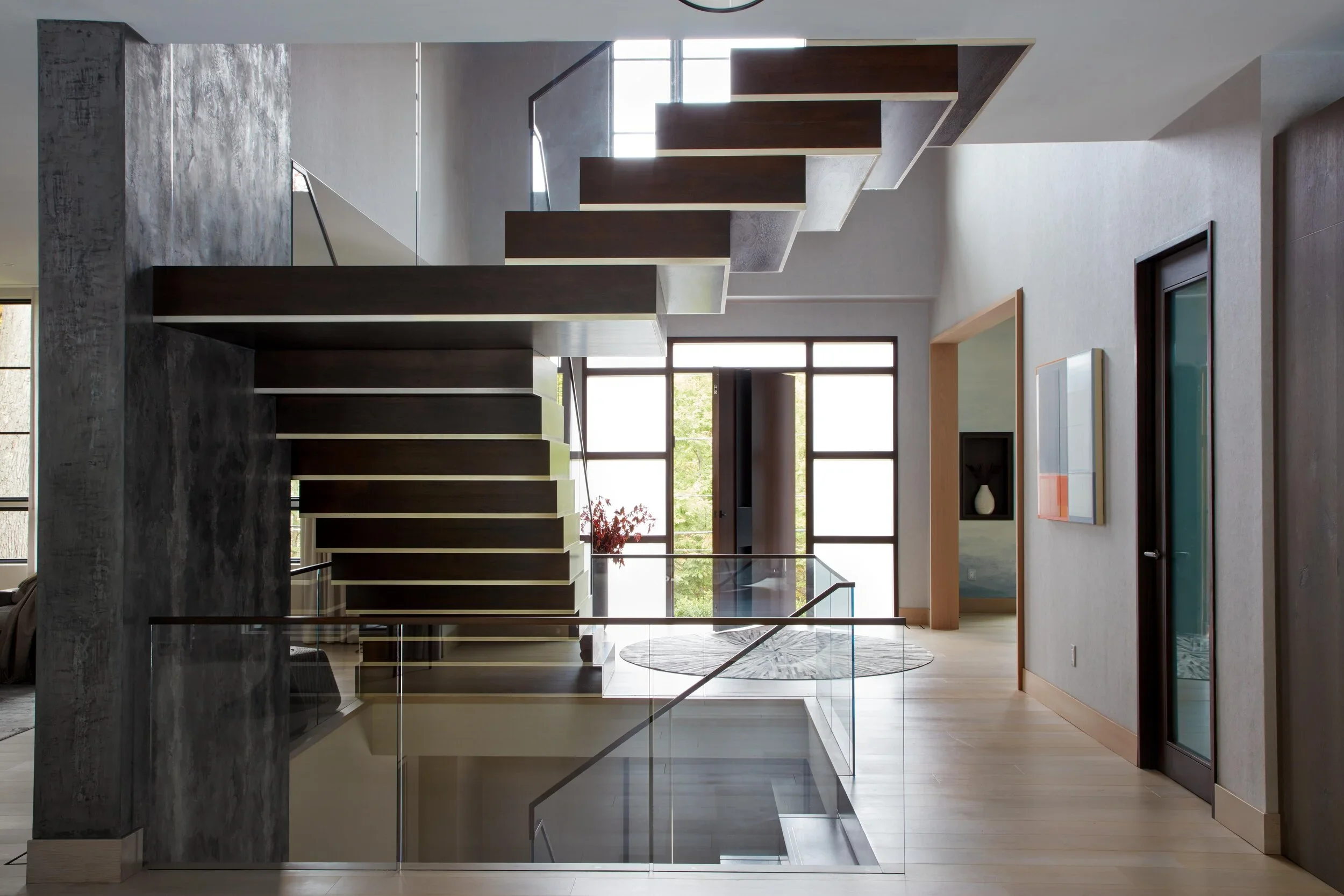 Interior of a modern home featuring a staircase with floating dark wood steps, glass railing, and large windows letting in natural light, with a wooden floor and contemporary wall art.