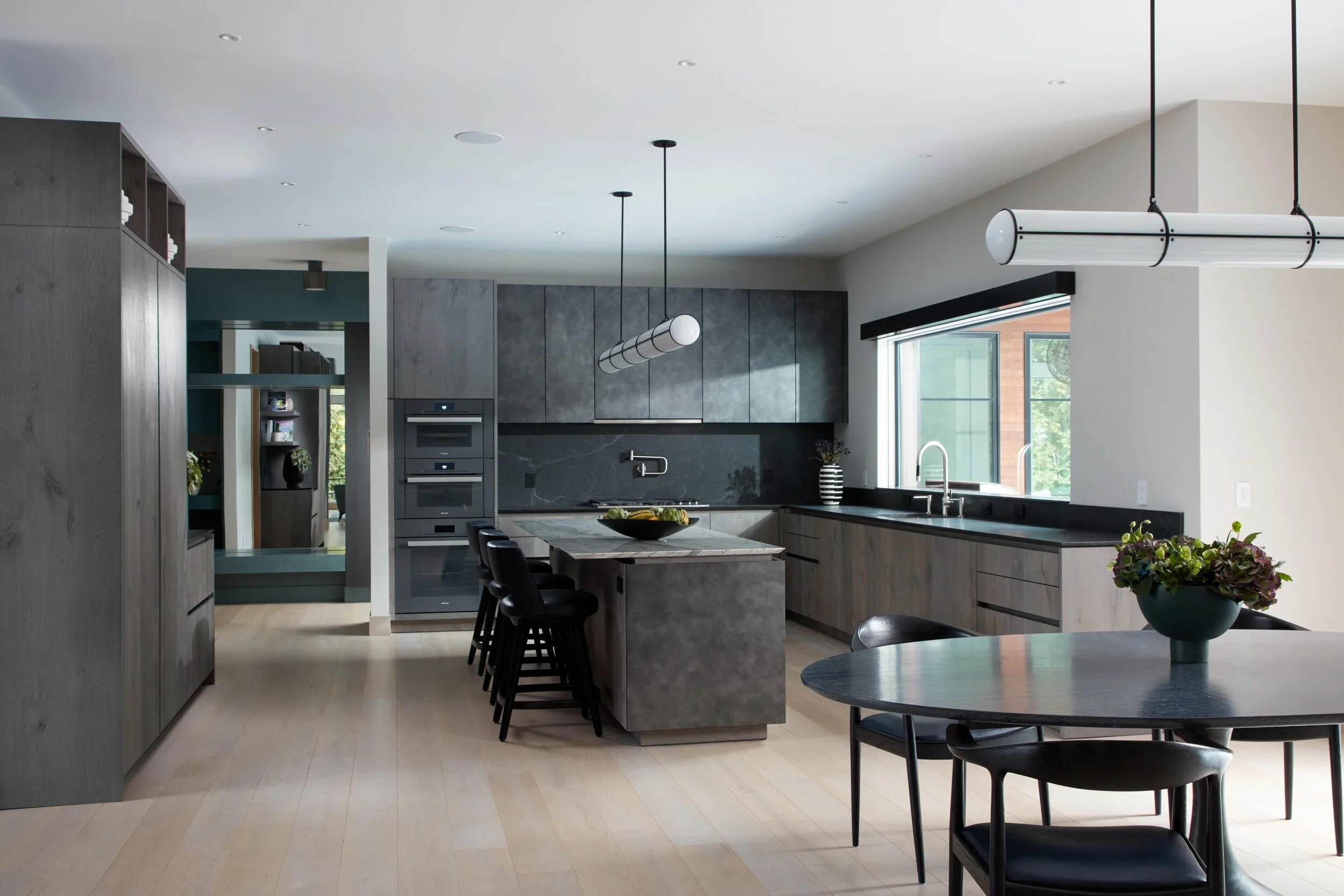 Modern kitchen with gray cabinets, black countertops, island with seating, sunlit window, and dining table with a flower vase.