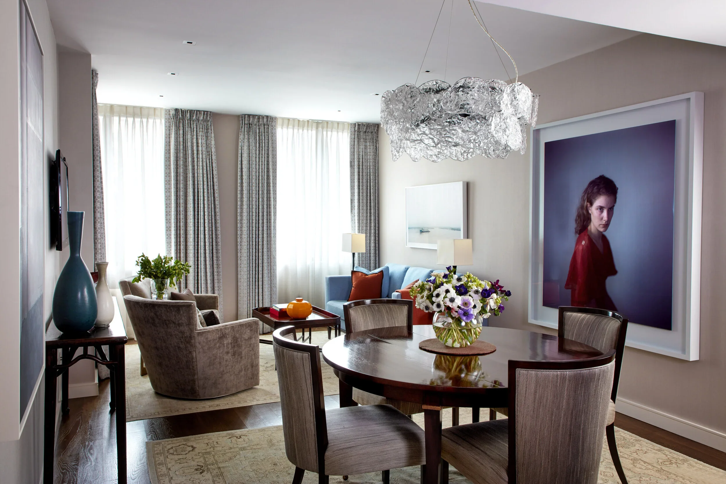 Living room with a round dining table with a flower arrangement, beige chairs, a blue sofa, a large framed photograph of a woman, and a chandelier, with curtains and a side table with vases.