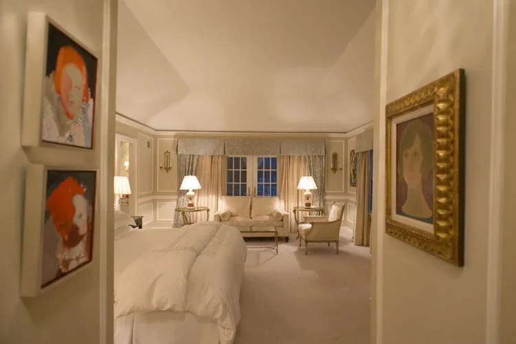 A luxurious bedroom with a white bed, white armchair, and beige sofa. The room has cream-colored walls, gold-framed paintings, and a window with light curtains and valance. Several lamps provide warm lighting.