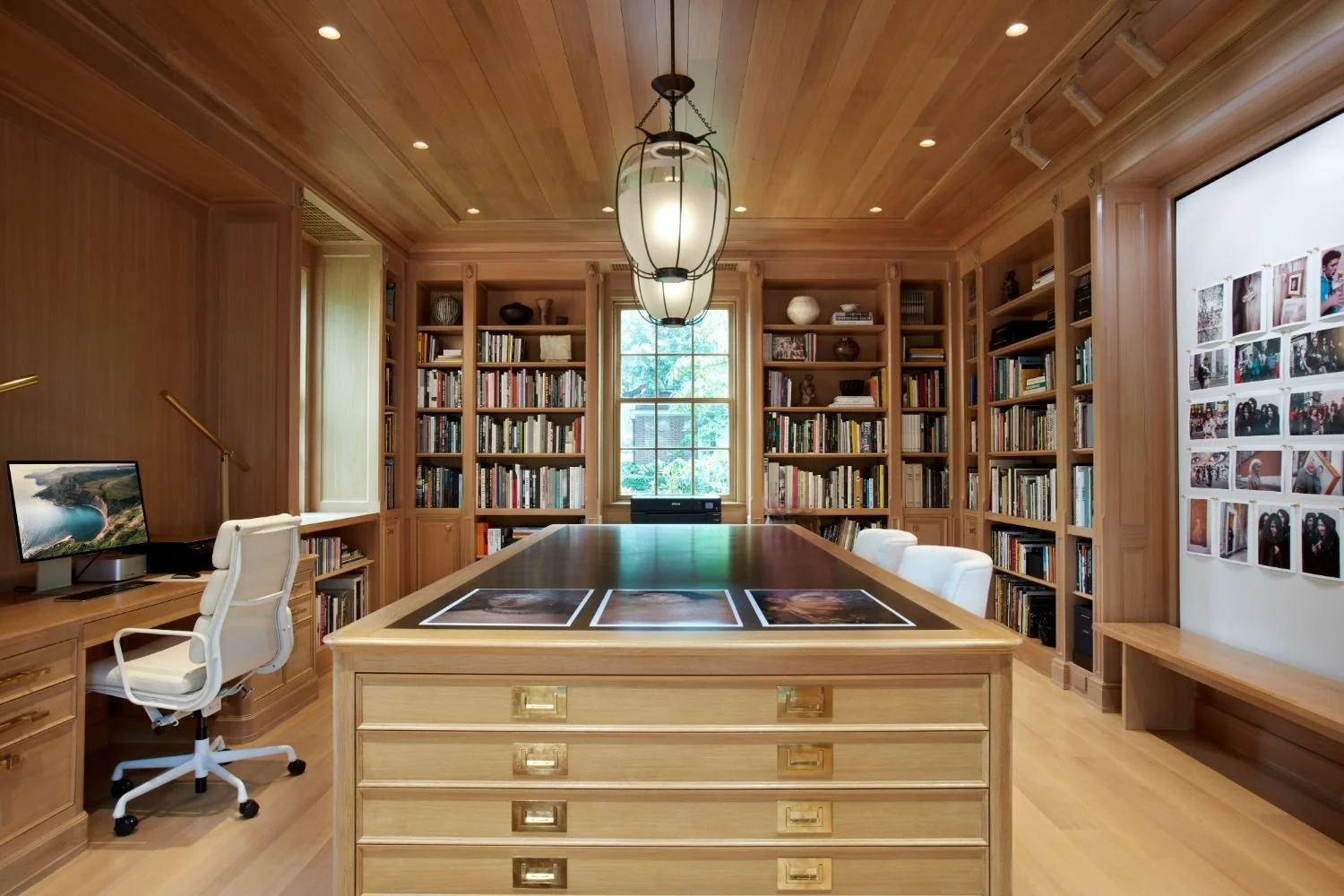 A home library with wooden walls, ceiling, and floors. Contains a large central wooden desk with photographs on top, white office chair, bookshelves filled with books, a window at the back, and a white wall decorated with photos.