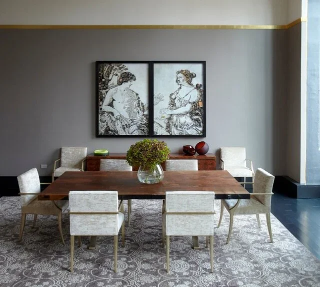 A dining room with a wooden table and eight upholstered chairs, a vase with green foliage on the table, a sideboard with decorative bowls and plants, and two framed monochrome artworks hanging on a gray wall behind the sideboard.