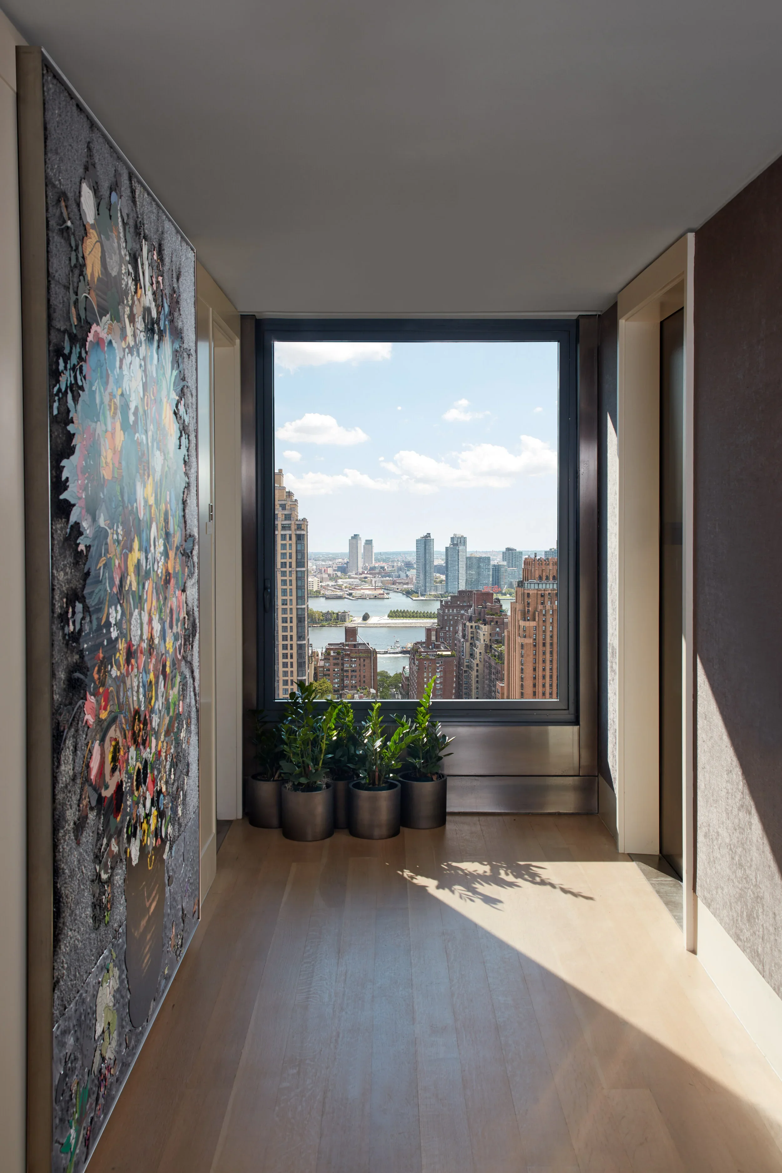 A corridor with a large window showing a cityscape view, potted plants on the floor, and a colorful abstract painting hanging on the left wall.