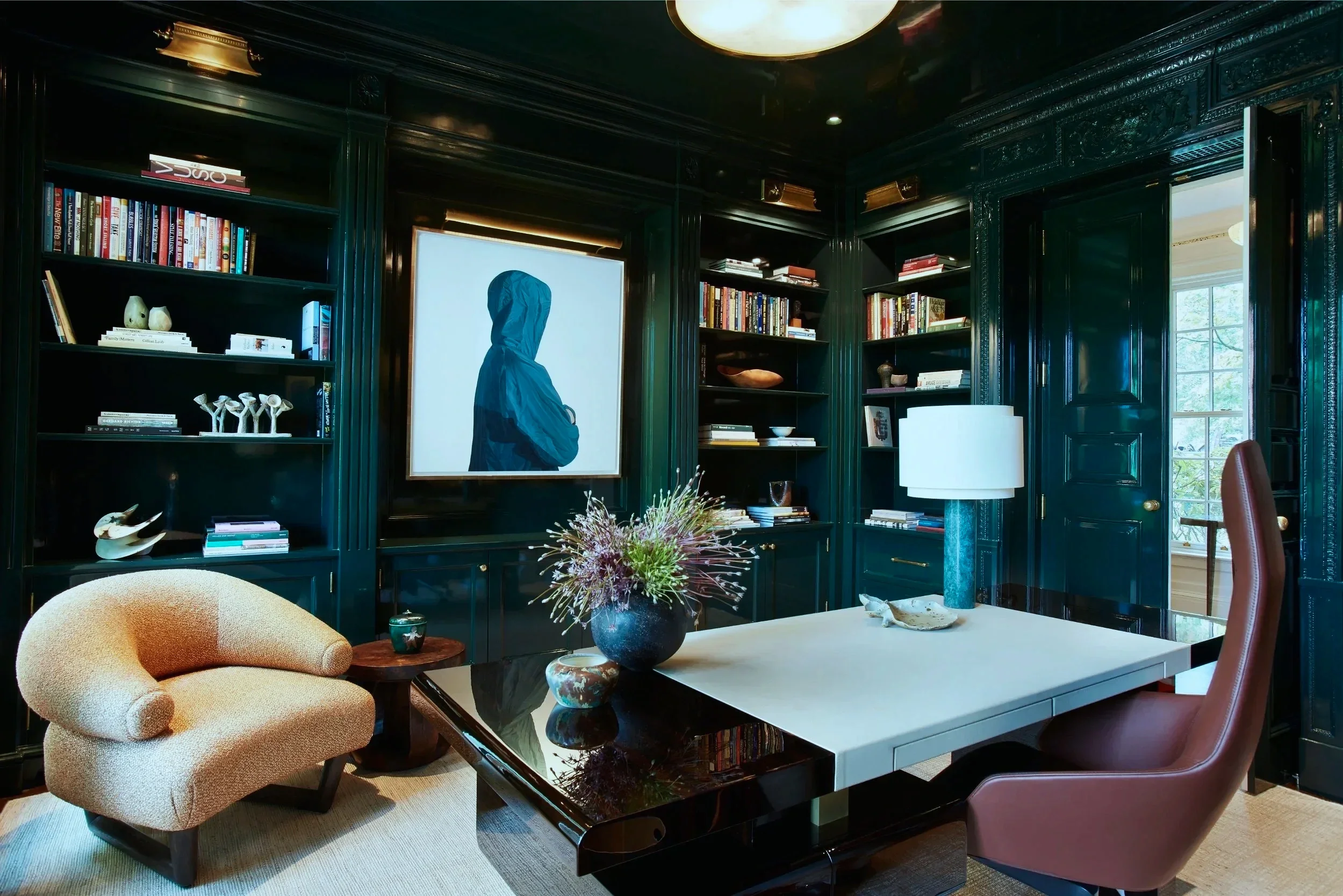 A luxurious home office with dark green paneled walls, built-in bookshelves filled with books and decorative objects, a large window letting in natural light, a white desk with a pink chair, a beige textured armchair, a side table with a cup, a table lamp, a vase with flowers, and a framed artwork of a person in a hooded jacket.