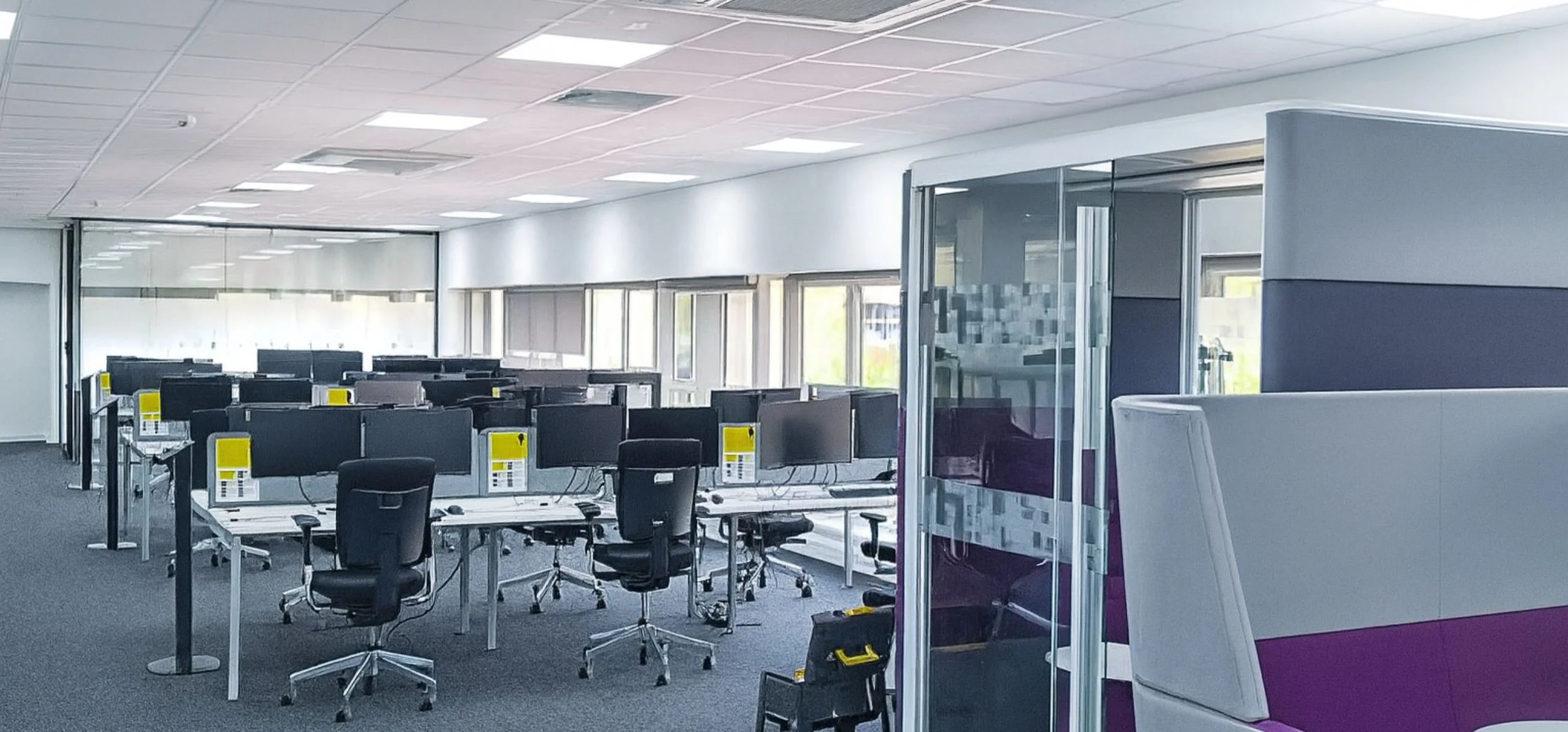 An empty office with multiple computer workstations, black chairs, and large windows letting in natural light.
