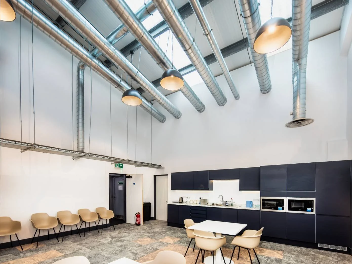 A modern industrial-style kitchen or break area with black cabinets, beige chairs, and air duct pipes on the ceiling.