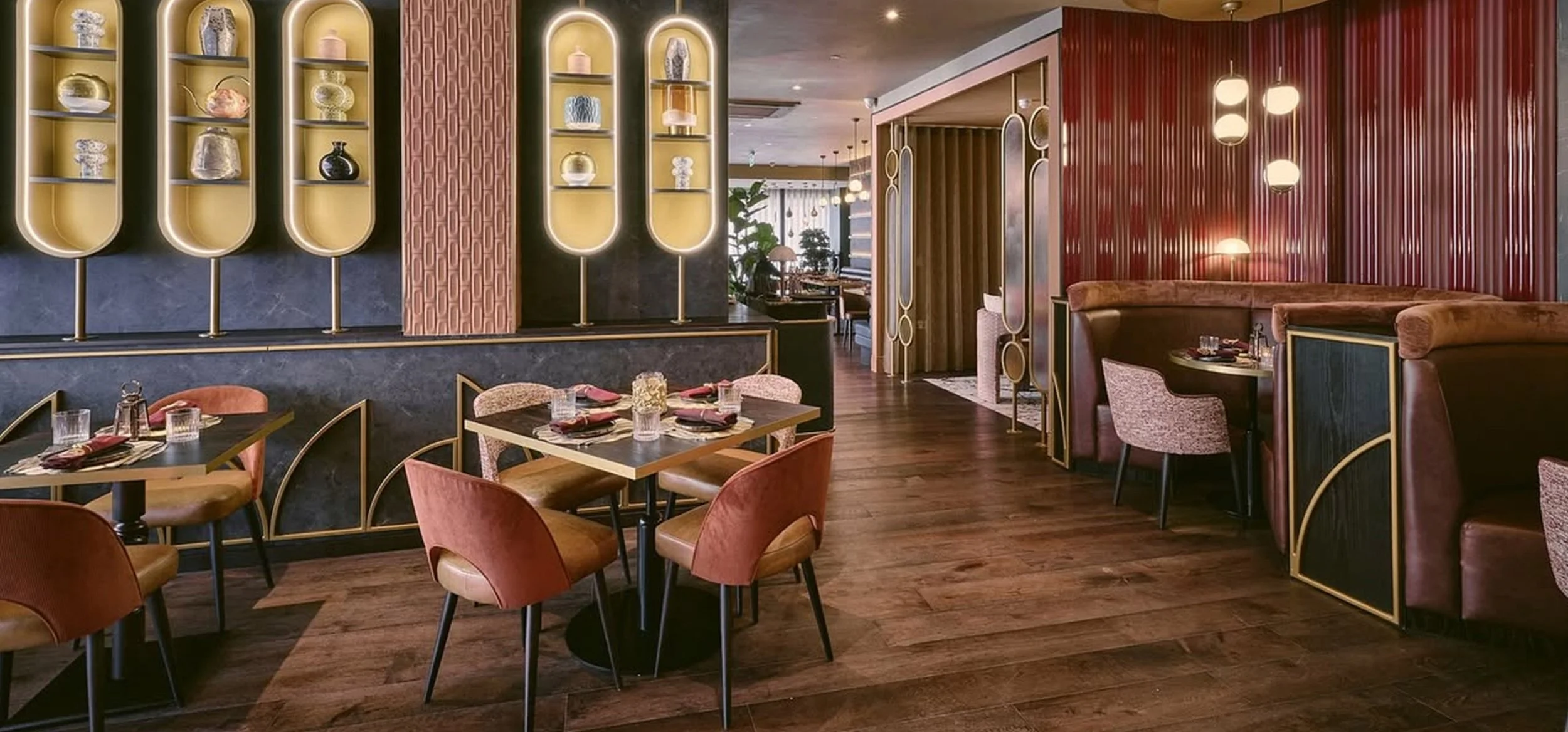 Elegant restaurant interior with dark wood flooring, pink and beige chairs, and a wall of decorative glass shelves displaying vases and art pieces. A semicircular booth with pink upholstery is visible on the right, and there are hanging pendant light