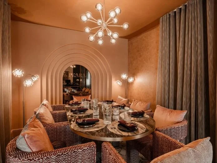 A luxurious dining room with a round marble table set with black plates, napkins, and glasses, surrounded by wicker chairs with pink cushions. The room has warm beige and peach walls, textured curtains, and modern spherical light fixtures on each side and a chandelier overhead, with an arched doorway in the background.