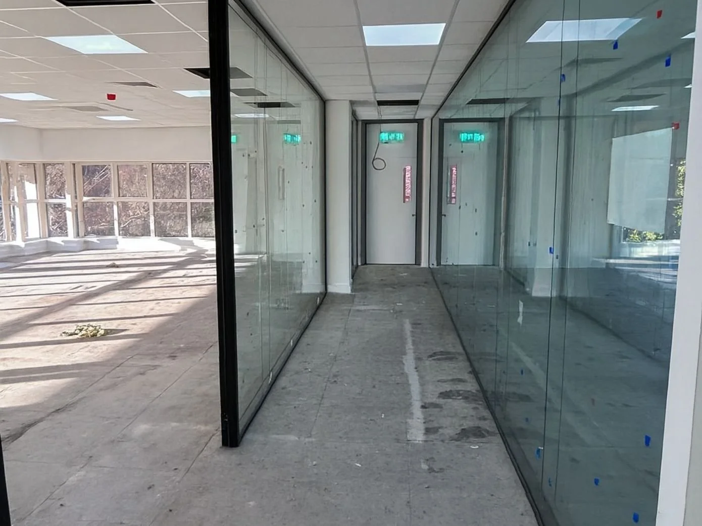 An indoor hallway with unfinished flooring, glass walls, and an exit door at the end. Lots of natural light coming through large windows, and some ceiling panels are missing.