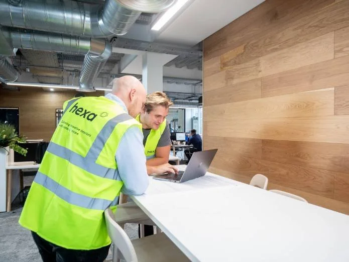 Two men wearing high-visibility vests, branded with 'Hexo,' working together on a laptop in a modern office with open ceiling pipes and wood-paneled walls.