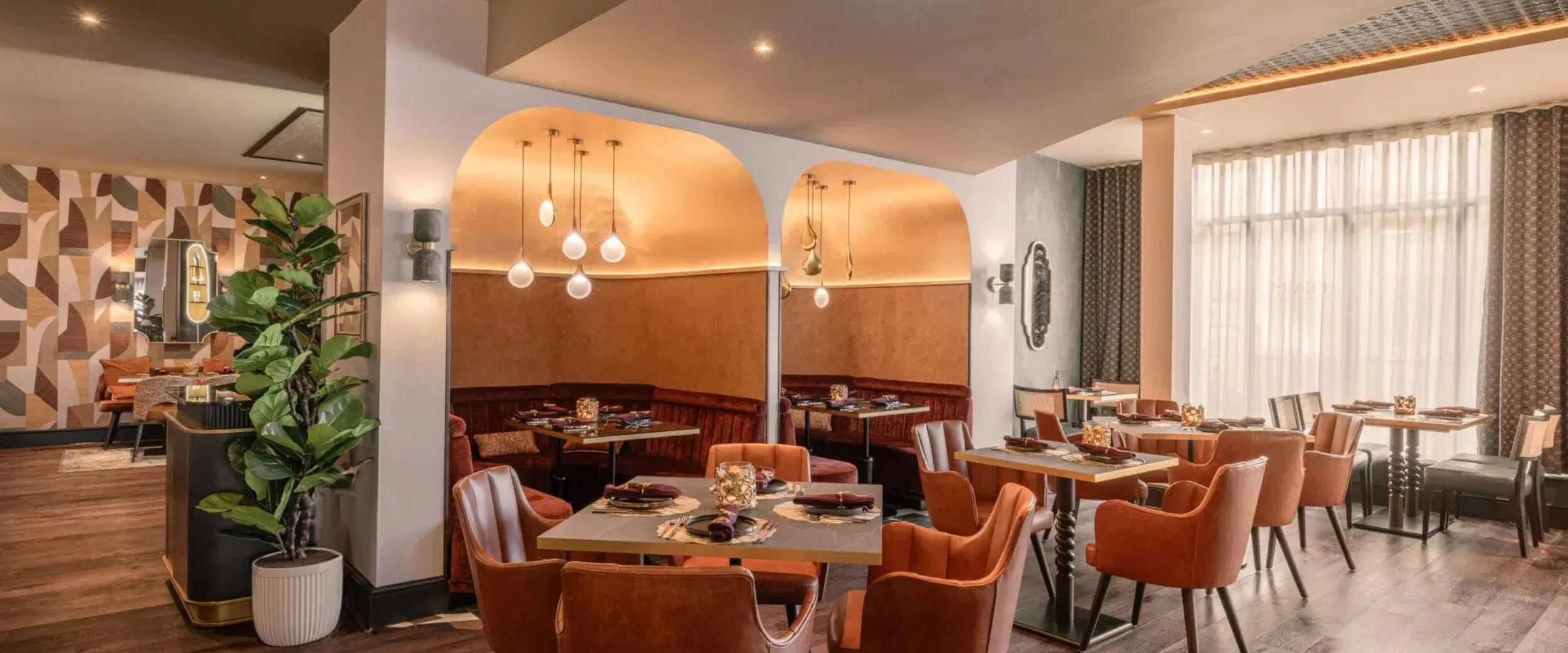 Interior of a modern restaurant with tables and chairs, orange velvet seating booths, decorative lighting, patterned curtains, potted plants, and artwork on the walls.