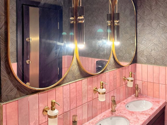 A pink marble double bathroom vanity with two sinks, gold faucets, and three cylindrical wall-mounted soap dispensers. Above the vanity are three large round mirrors with gold frames, and black and gold light fixtures are mounted above each mirror. T