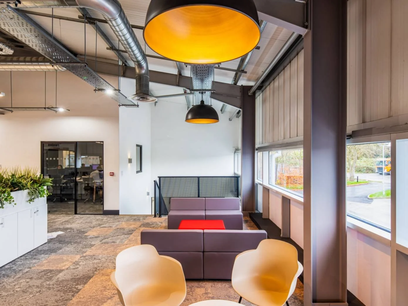 Modern office lounge area with beige chairs, a purple sofa with a red cushion, large windows with view of parking lot, black and yellow hanging lamps, and an open ceiling with industrial ductwork.
